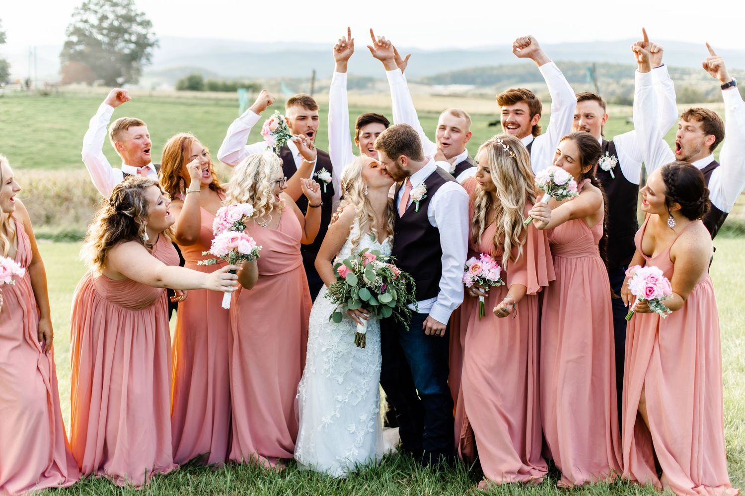 Joyful wedding party celebrating with bride and groom in field with pink bridesmaid dresses.