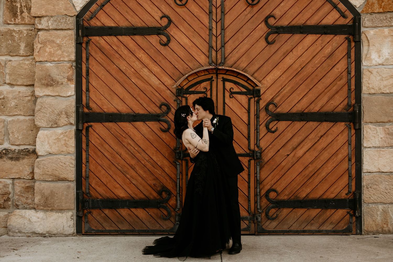 Bride in black gown and groom kissing in front of rustic wooden doors at wedding.