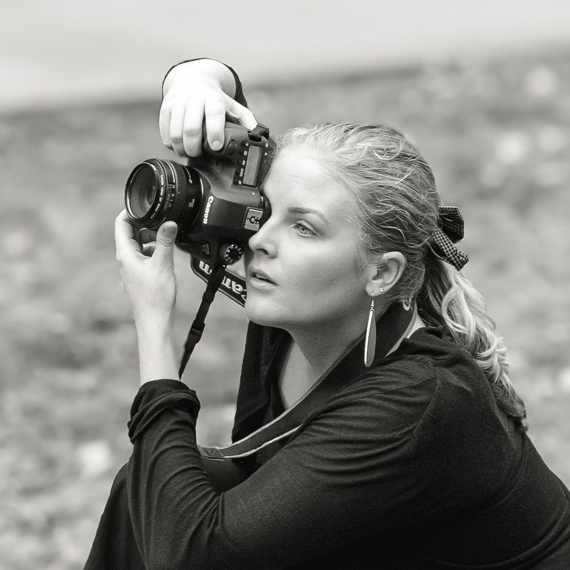 Close-up black and white portrait of blonde woman holding DSLR camera up to take a photo outdoors.