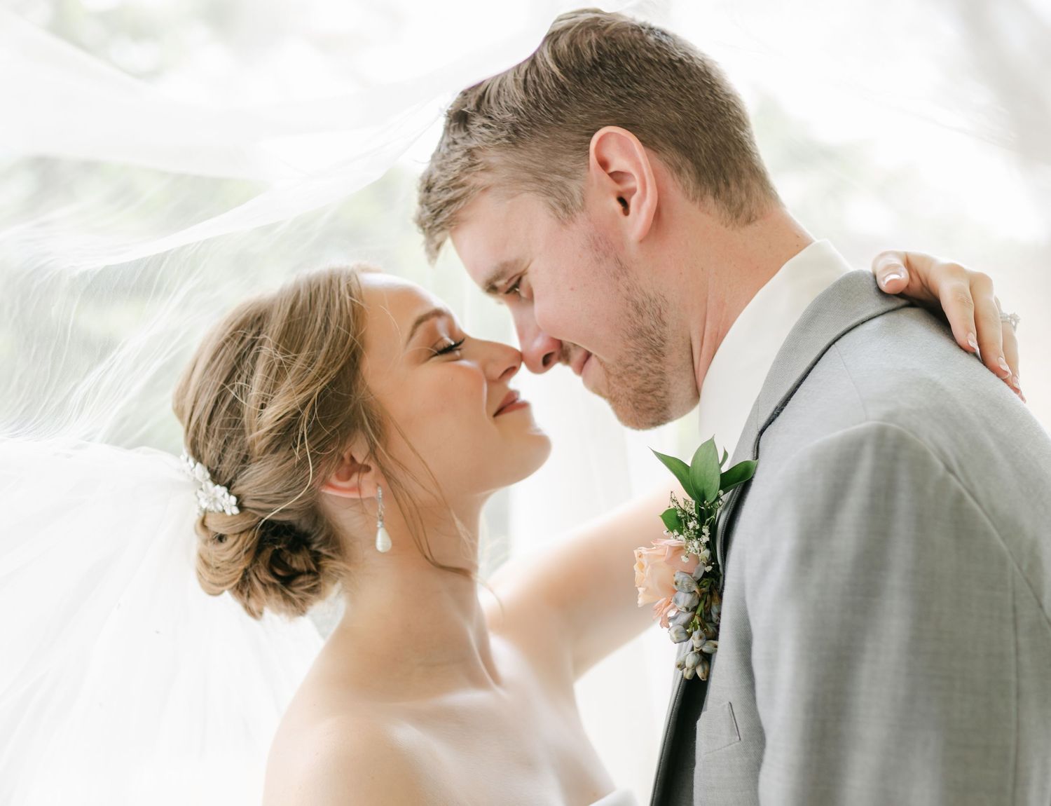 Bride and groom share tender almost-kiss moment under billowing white veil outdoors on wedding day.