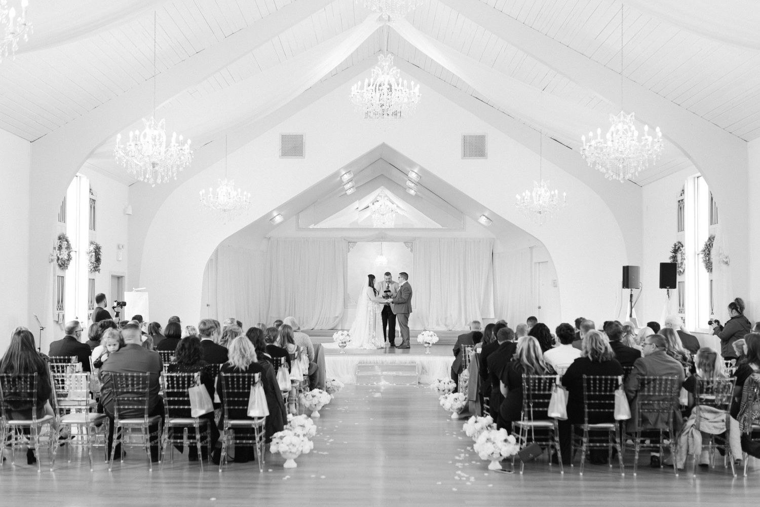 Black and white photo of grand wedding ceremony in elegant white draped venue with chandeliers.