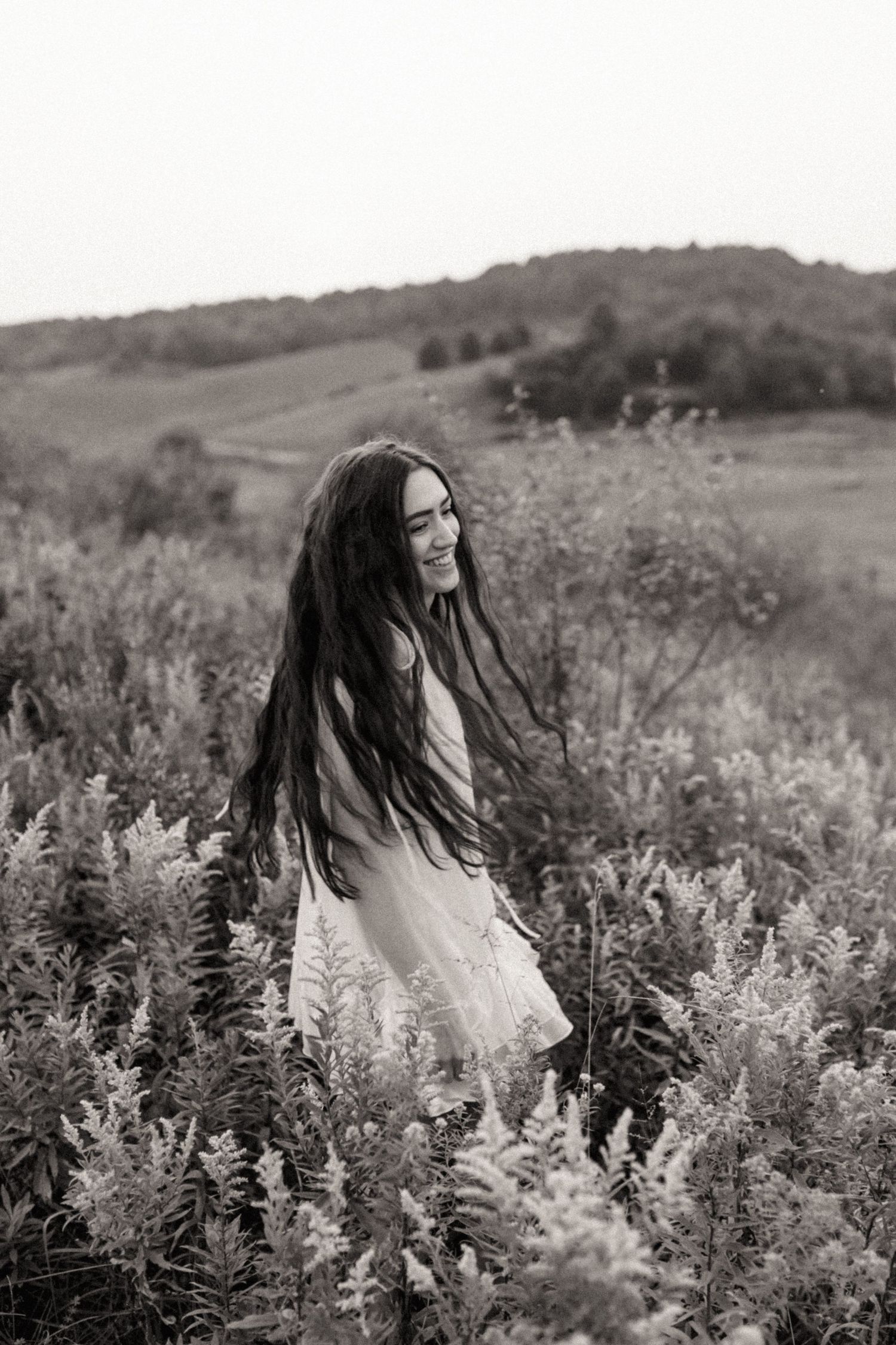 Black and white portrait of smiling woman with flowing hair in wildflower field.