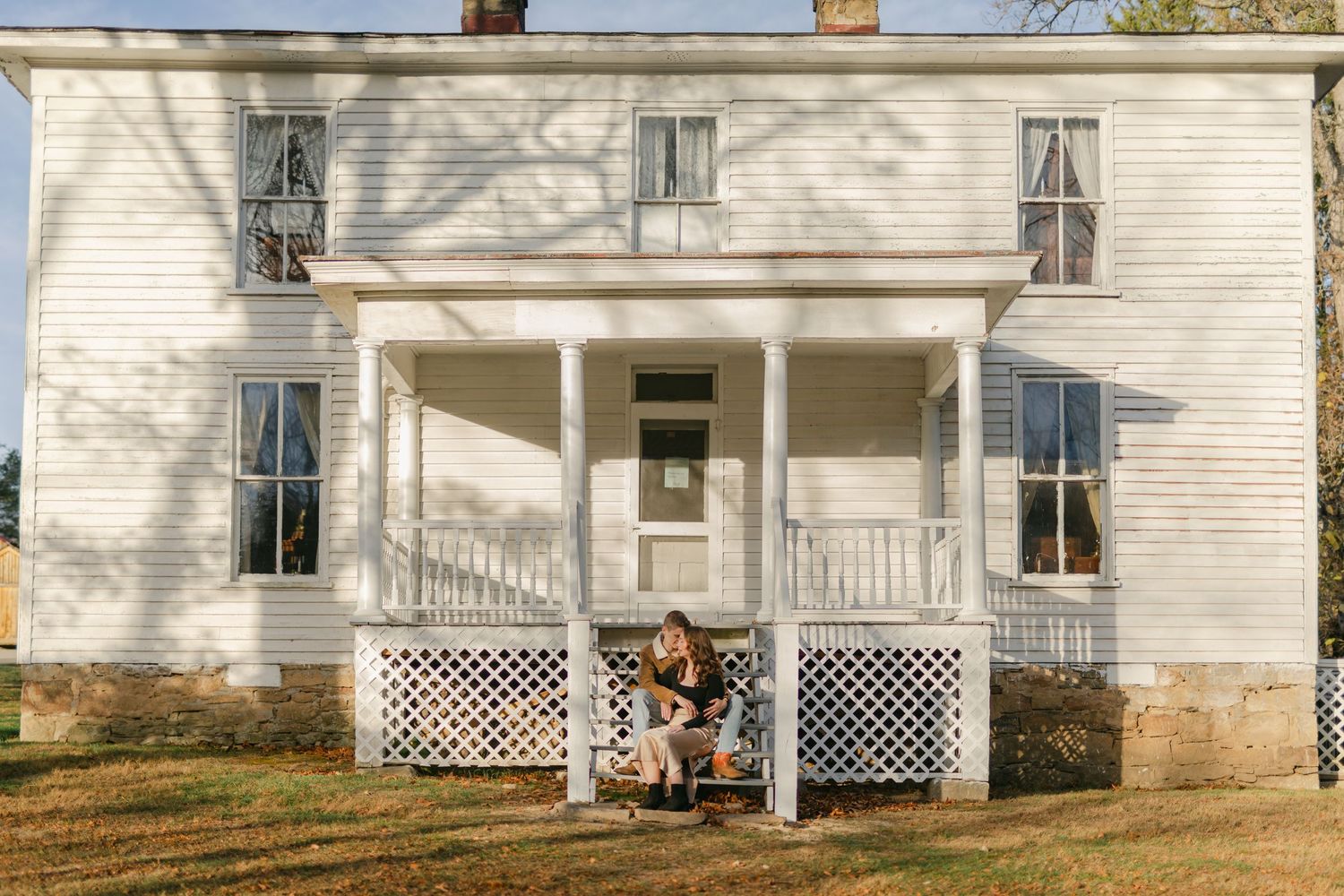 Person sitting on steps of a charming white two-story farmhouse with a covered front porch.
