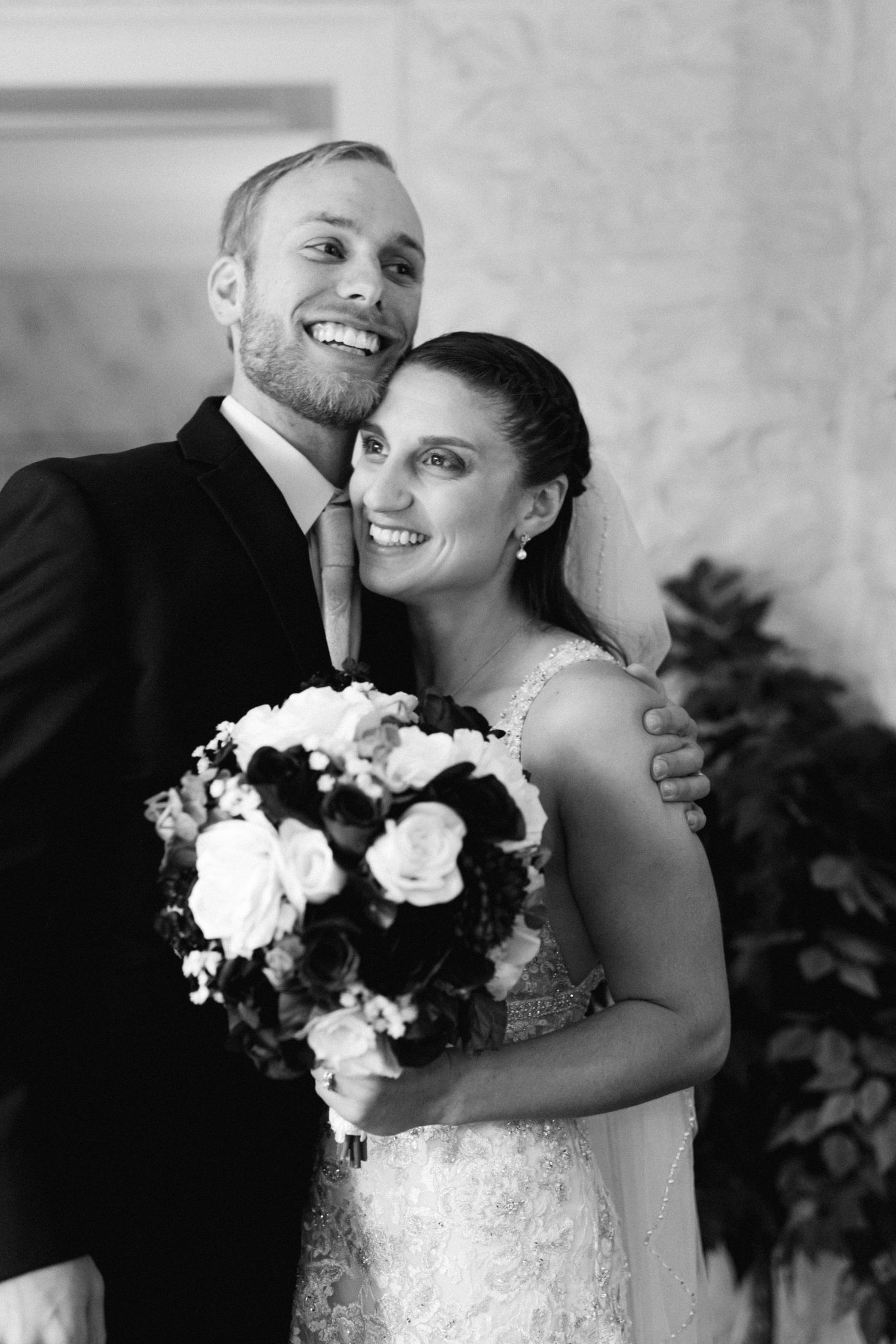 Black and white portrait of smiling bride and groom, bride holding elegant floral bouquet inside a classic venue.