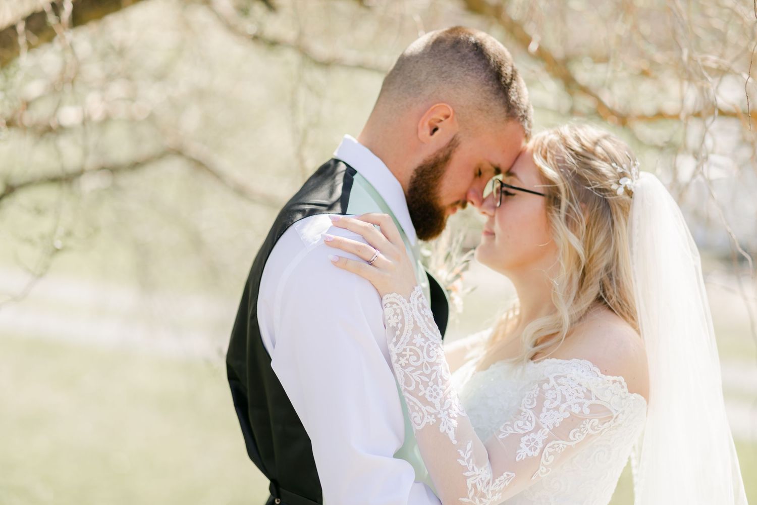 Bride and groom share an intimate forehead touch outdoors, bride in lace dress with veil, groom in black vest and tie.