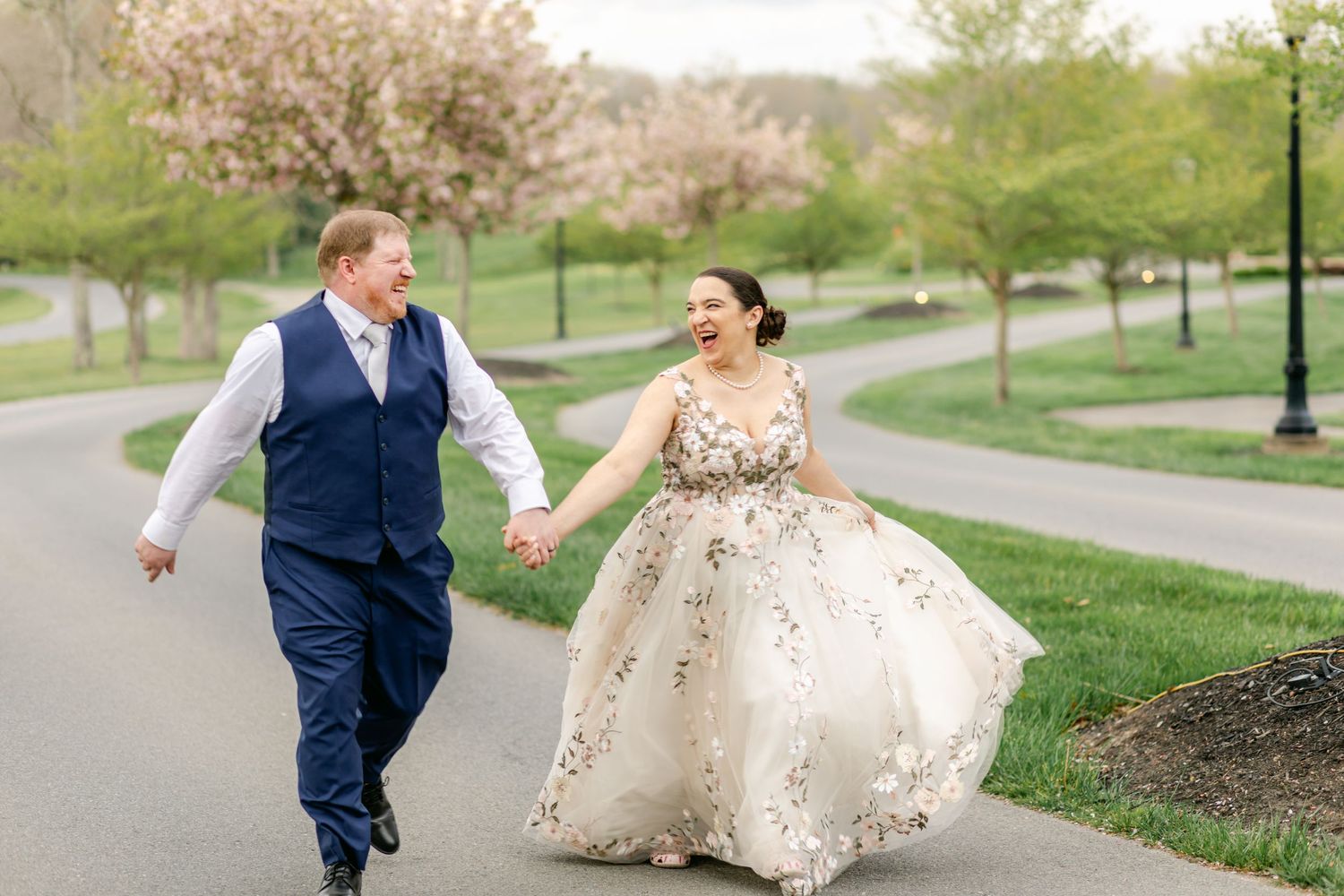 Joyful bride and groom running hand-in-hand on a path surrounded by blooming spring trees.