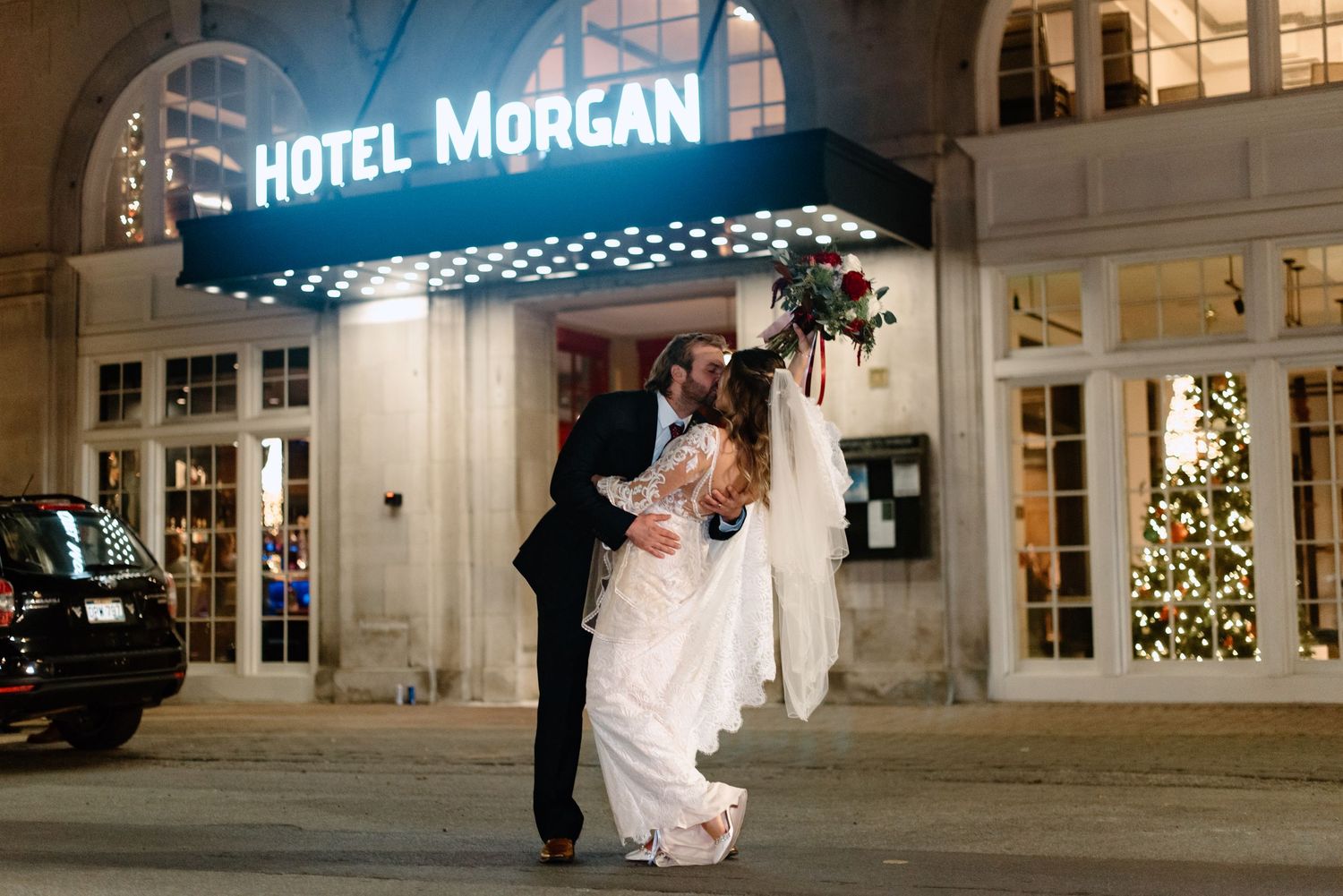 Bride and groom share a dramatic dip kiss outside Hotel Morgan at night, with festive Christmas decorations nearby.
