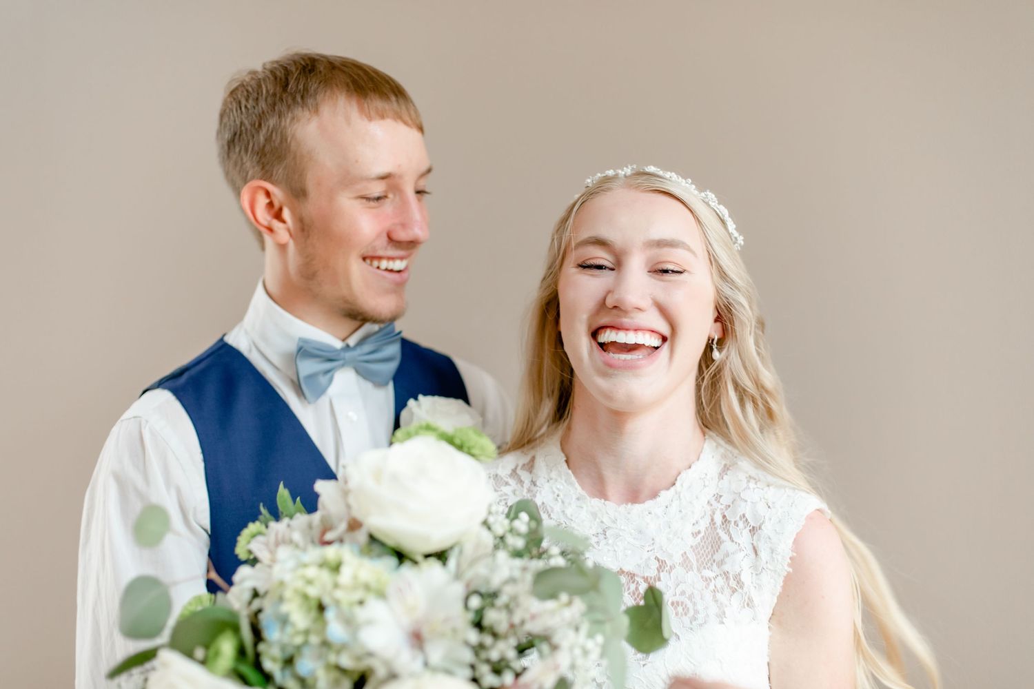 Laughing newlyweds posing together with a white rose bouquet against a neutral background.