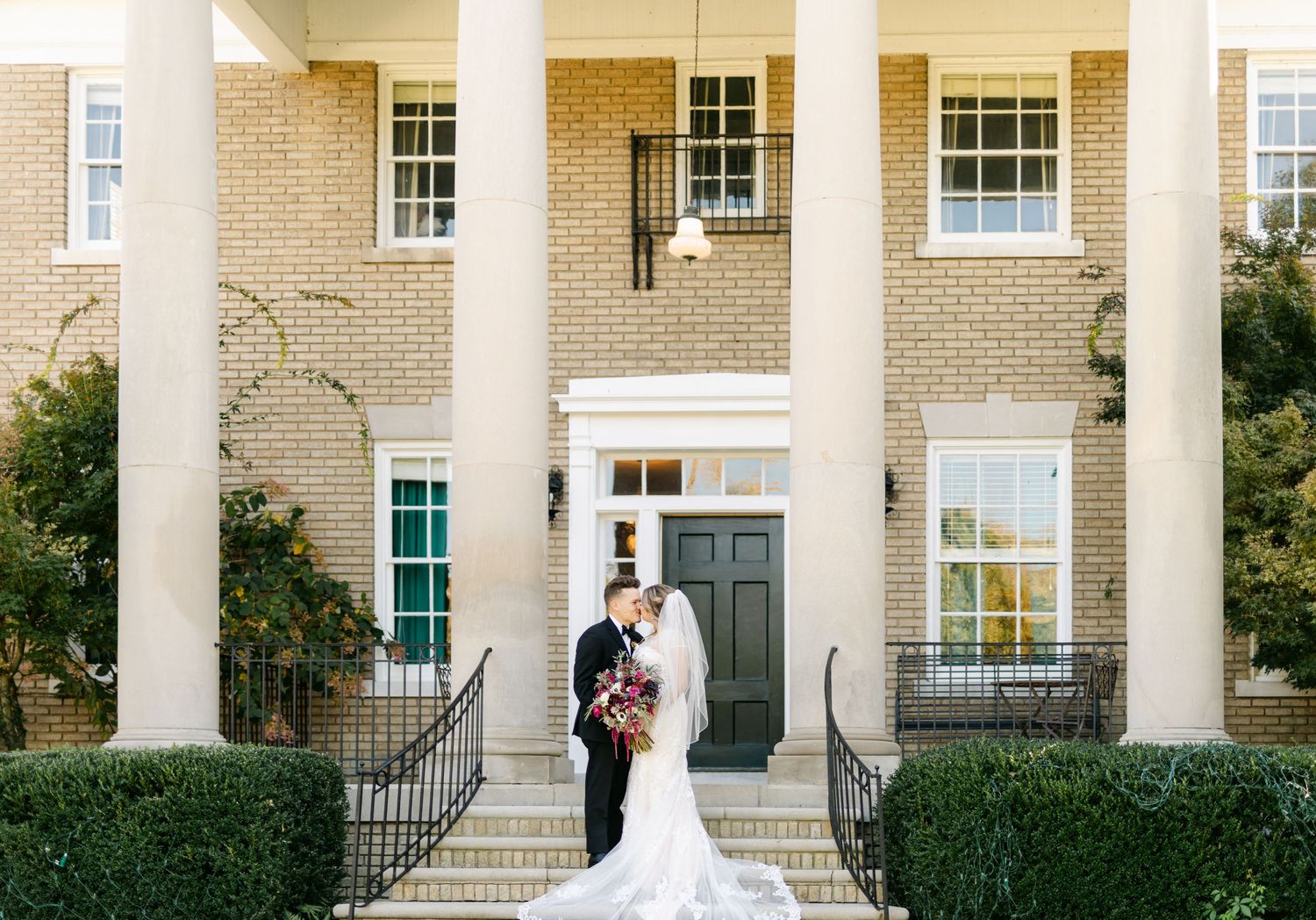 Bride and groom kissing on steps of elegant colonial-style wedding venue with columns.