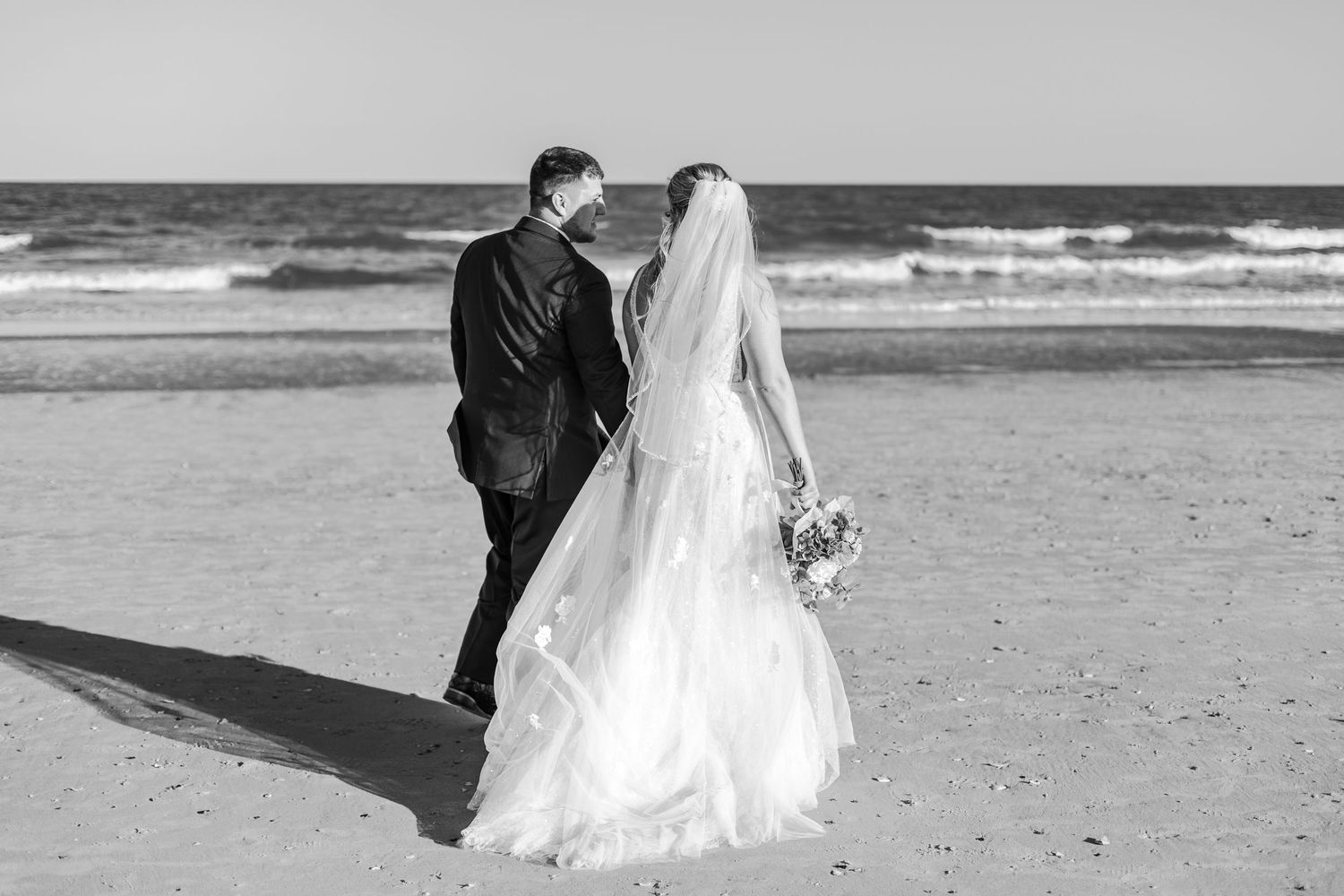 Black and white photo of bride and groom walking hand in hand along sandy beach.