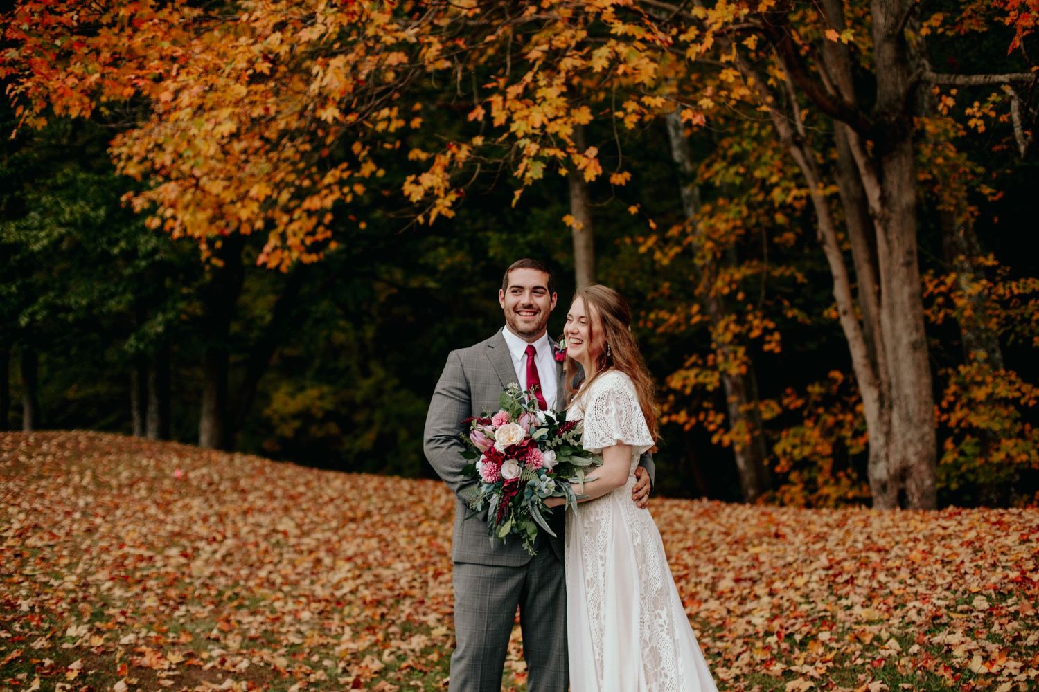 Smiling couple poses together in a stunning autumn forest setting with vibrant orange and red fallen leaves on the ground.