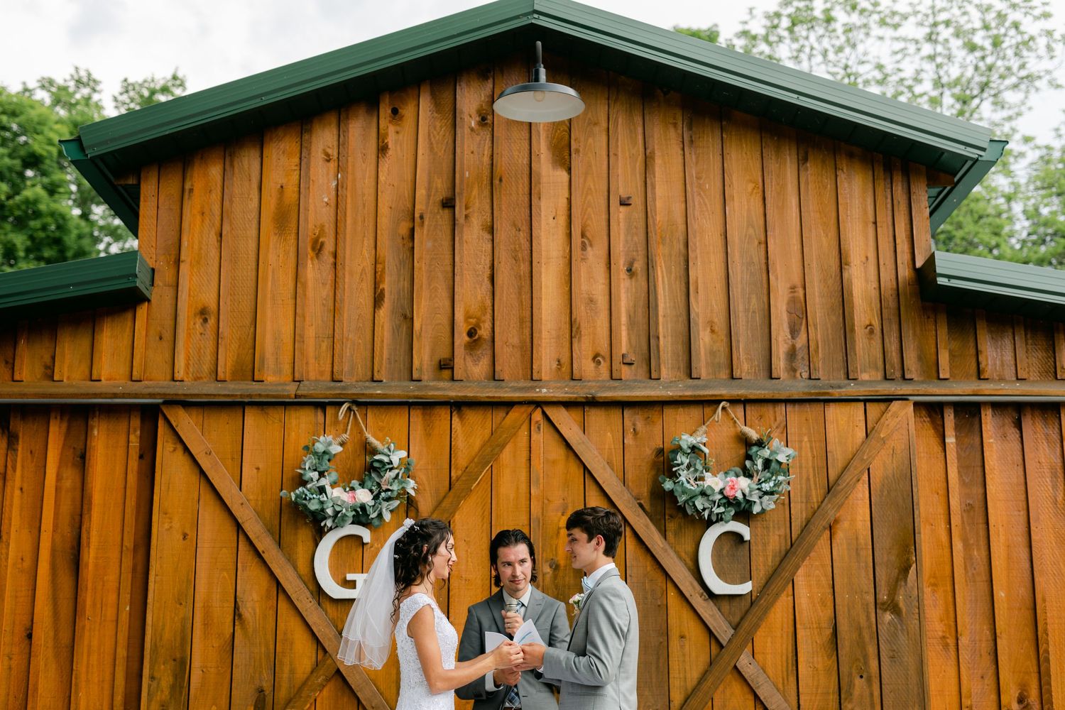 Bride and groom exchanging vows with officiant in front of rustic wooden barn doors with wreaths.