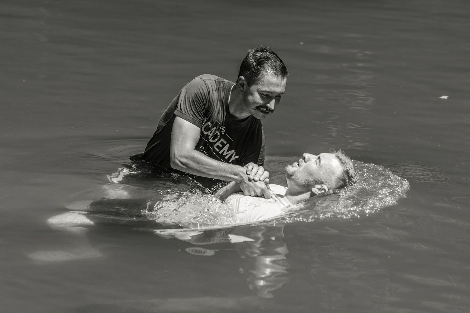 Man performing a baptism in a river, gently lowering a person into the water in a black and white photo.