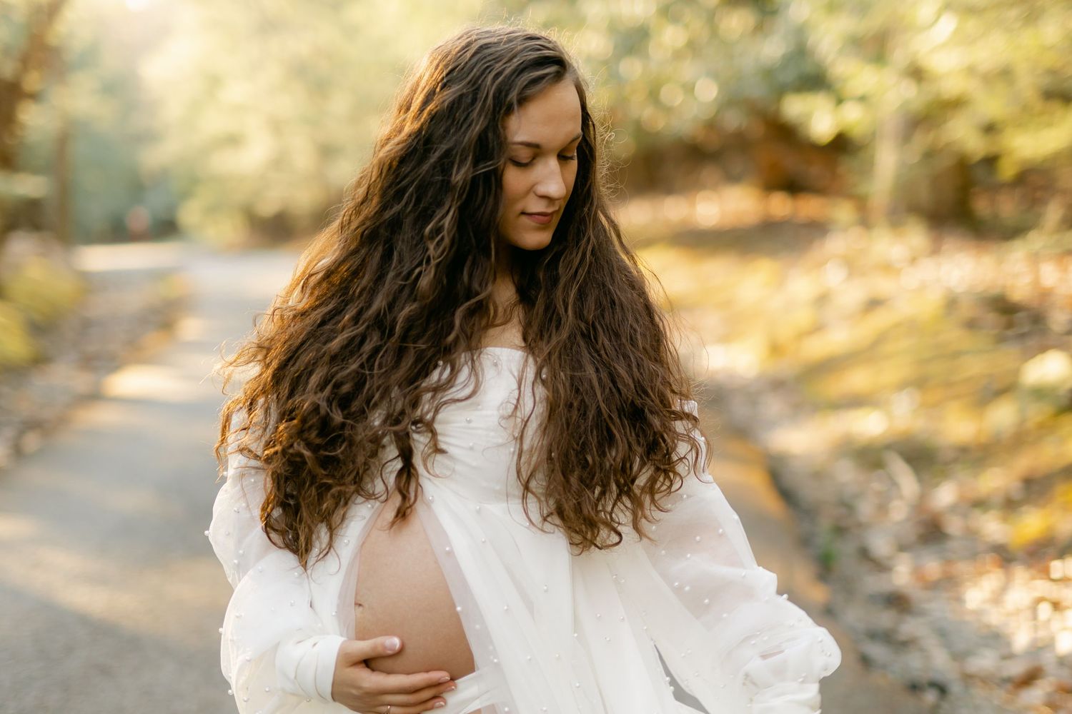 Pregnant woman in white dress holding baby bump outdoors in soft natural light.