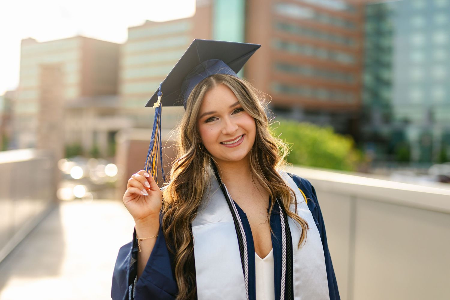 Smiling young woman in graduation cap and gown holding tassel outdoors in urban setting at sunset.