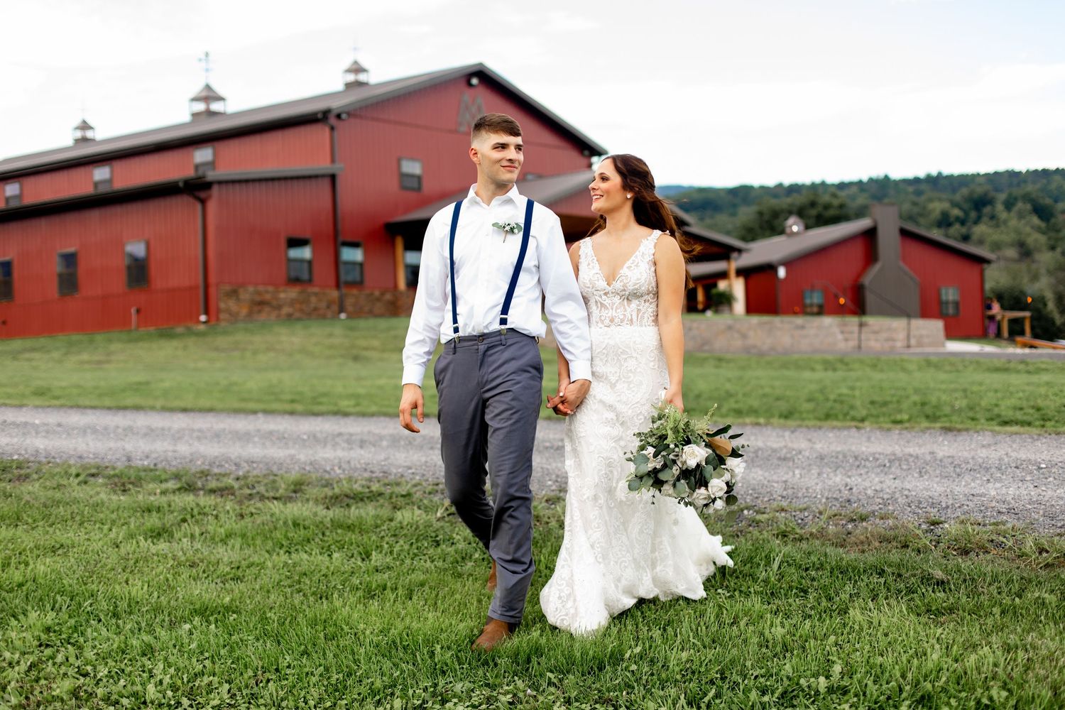 Bride and groom walking hand-in-hand near a red barn on a scenic countryside wedding venue.