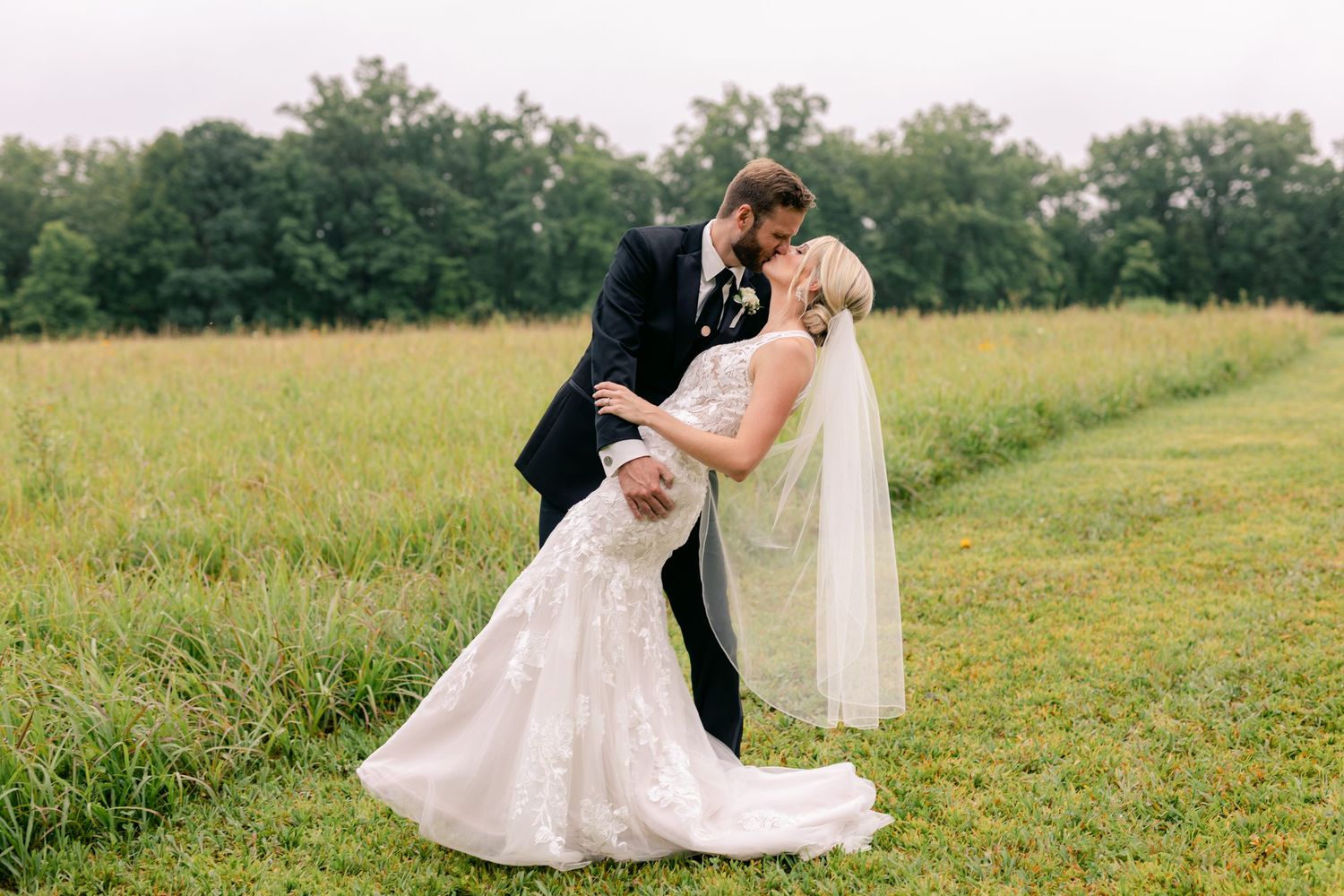 Bride and groom share romantic dip kiss in lush green field on their wedding day.