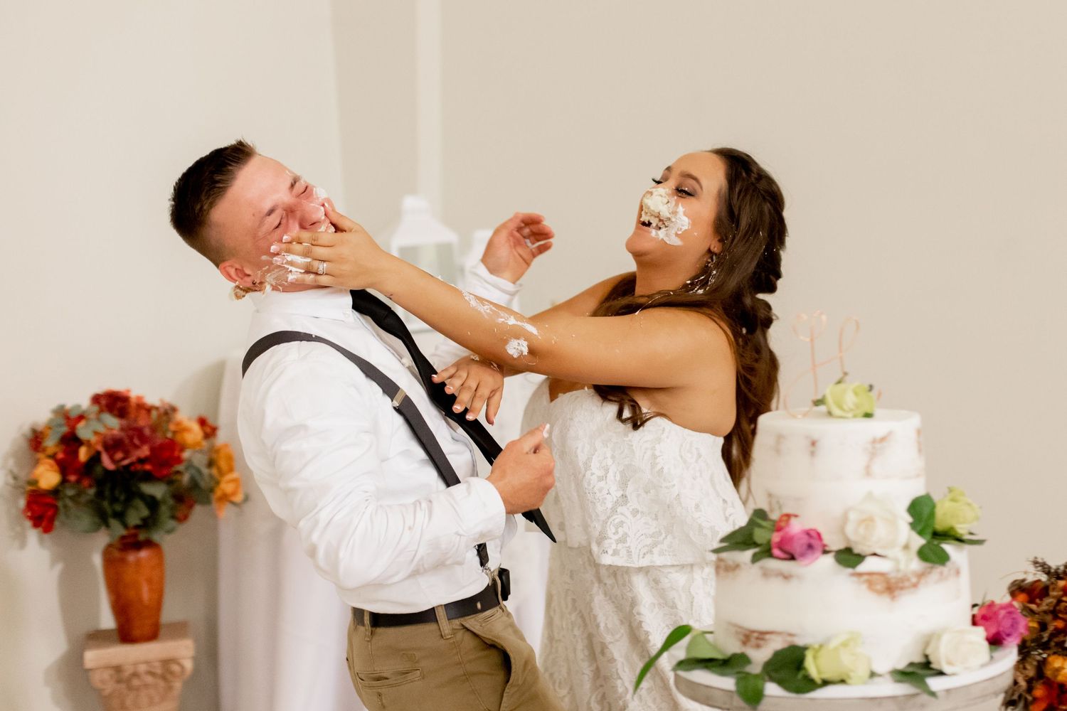 Playful newlyweds smashing wedding cake on each other faces beside a tiered white wedding cake.