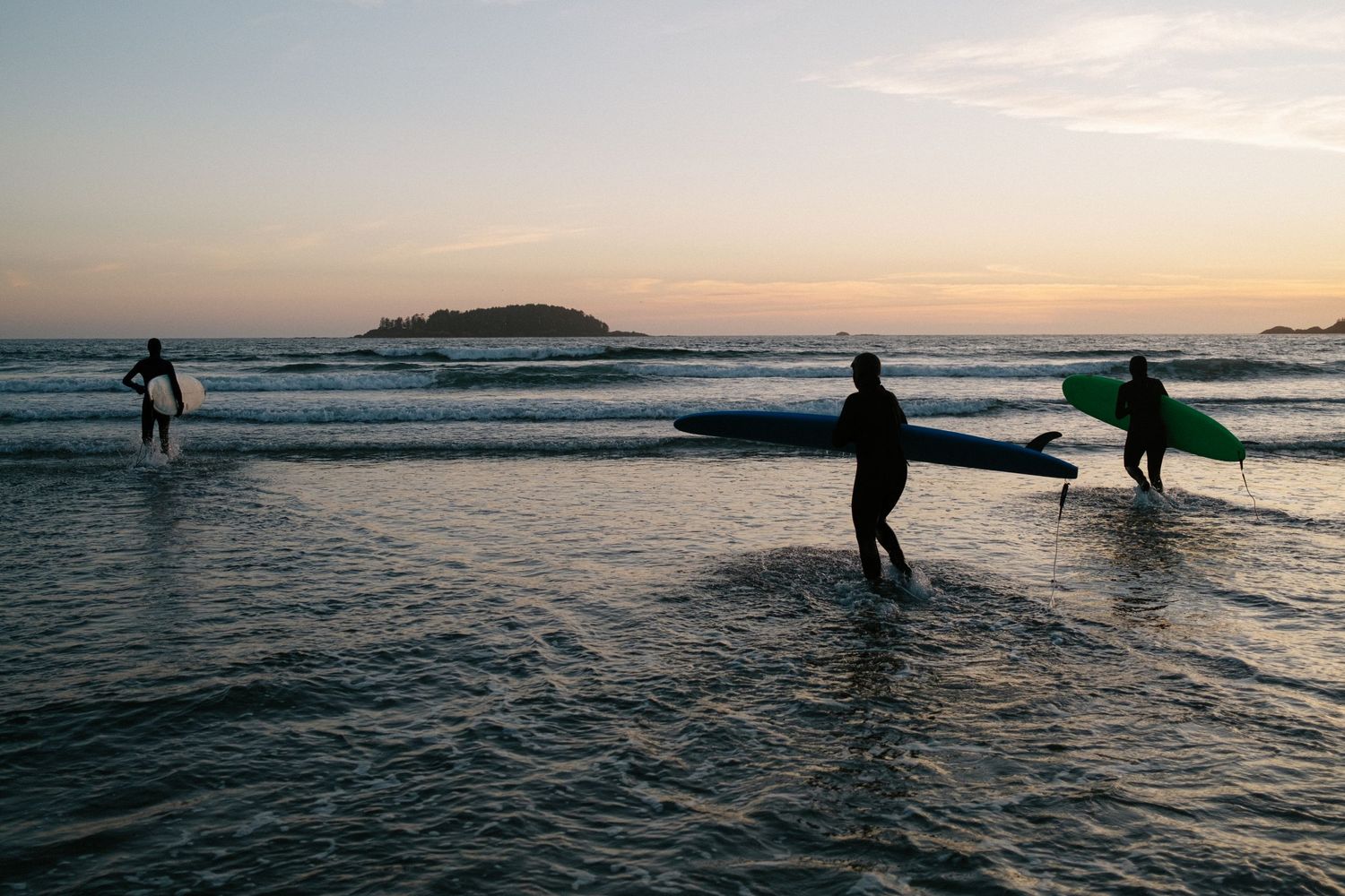 Silhouetted surfers walk along the shoreline during a golden sunset with an island in the background.