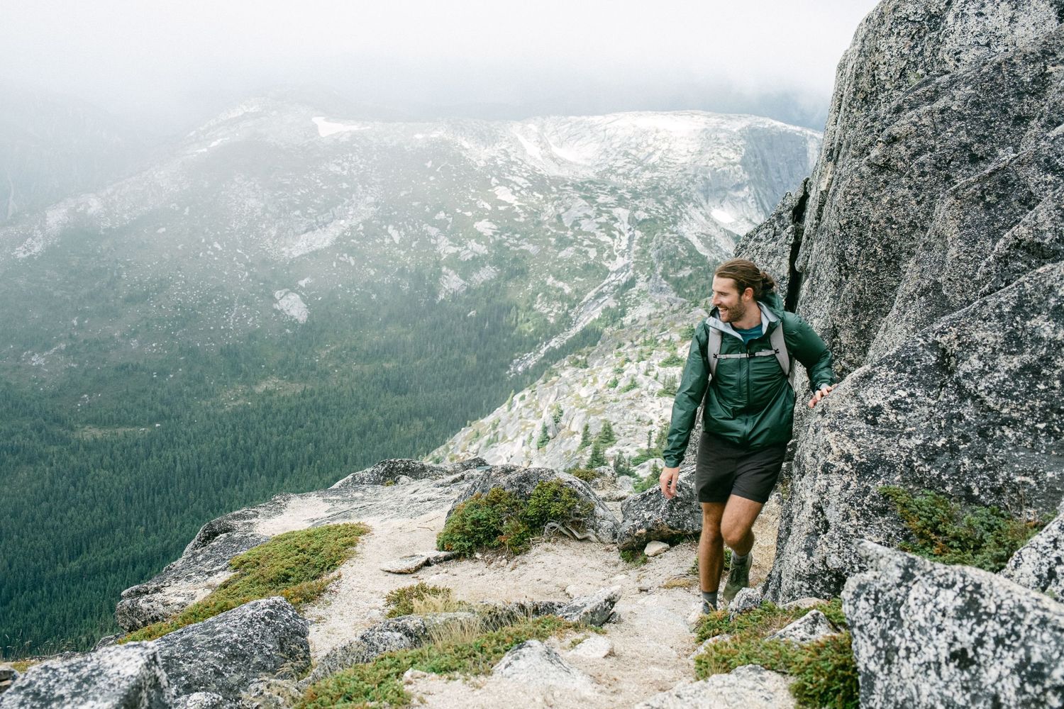 Hiker in green jacket traversing steep mountain trail with valley views below.