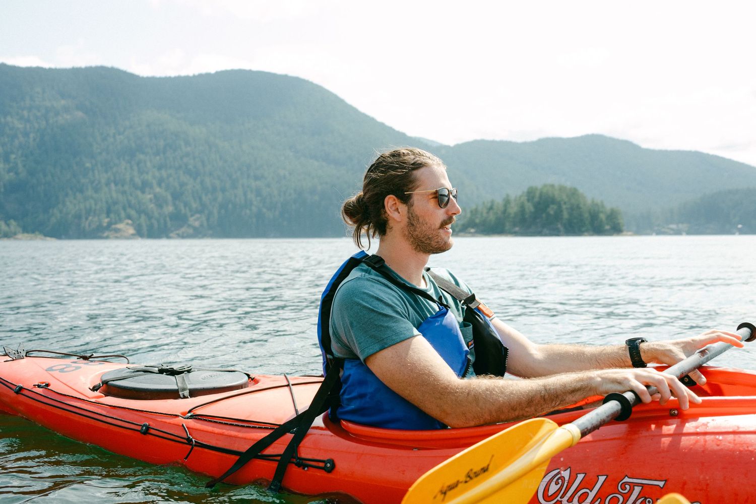 Kayaker paddling red kayak on calm lake surrounded by forested mountains.