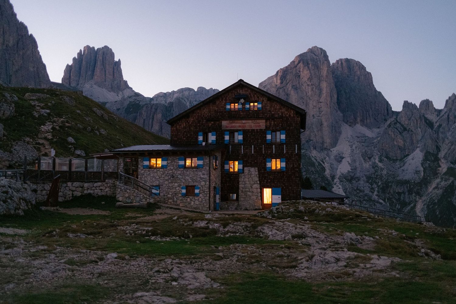 Rustic mountain lodge illuminated at dusk with dramatic Dolomites peaks towering in the background.