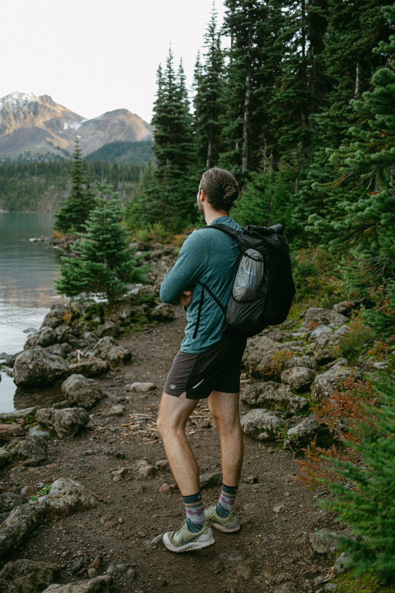 Hiker on rocky trail overlooking pristine mountain lake surrounded by evergreen forest.