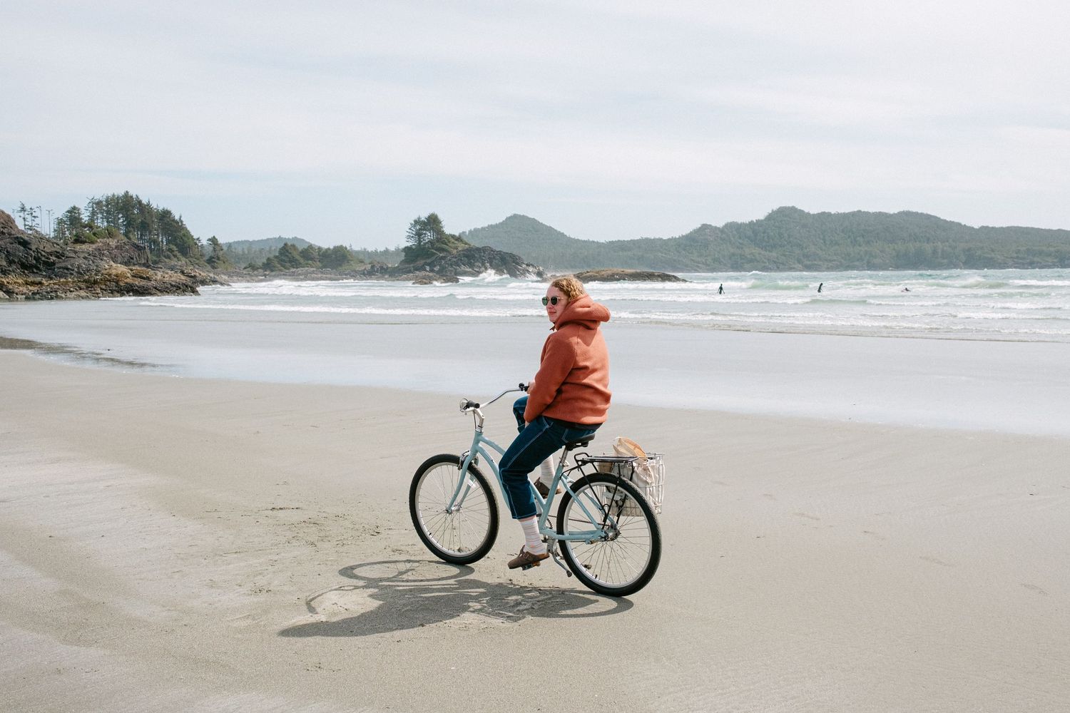 Person riding a bicycle along a scenic coastal beach with mountains in the distance.