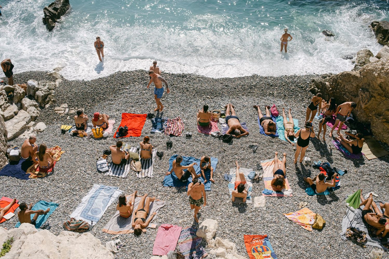 Aerial view of crowded rocky beach with sunbathers and swimmers enjoying Mediterranean waters.