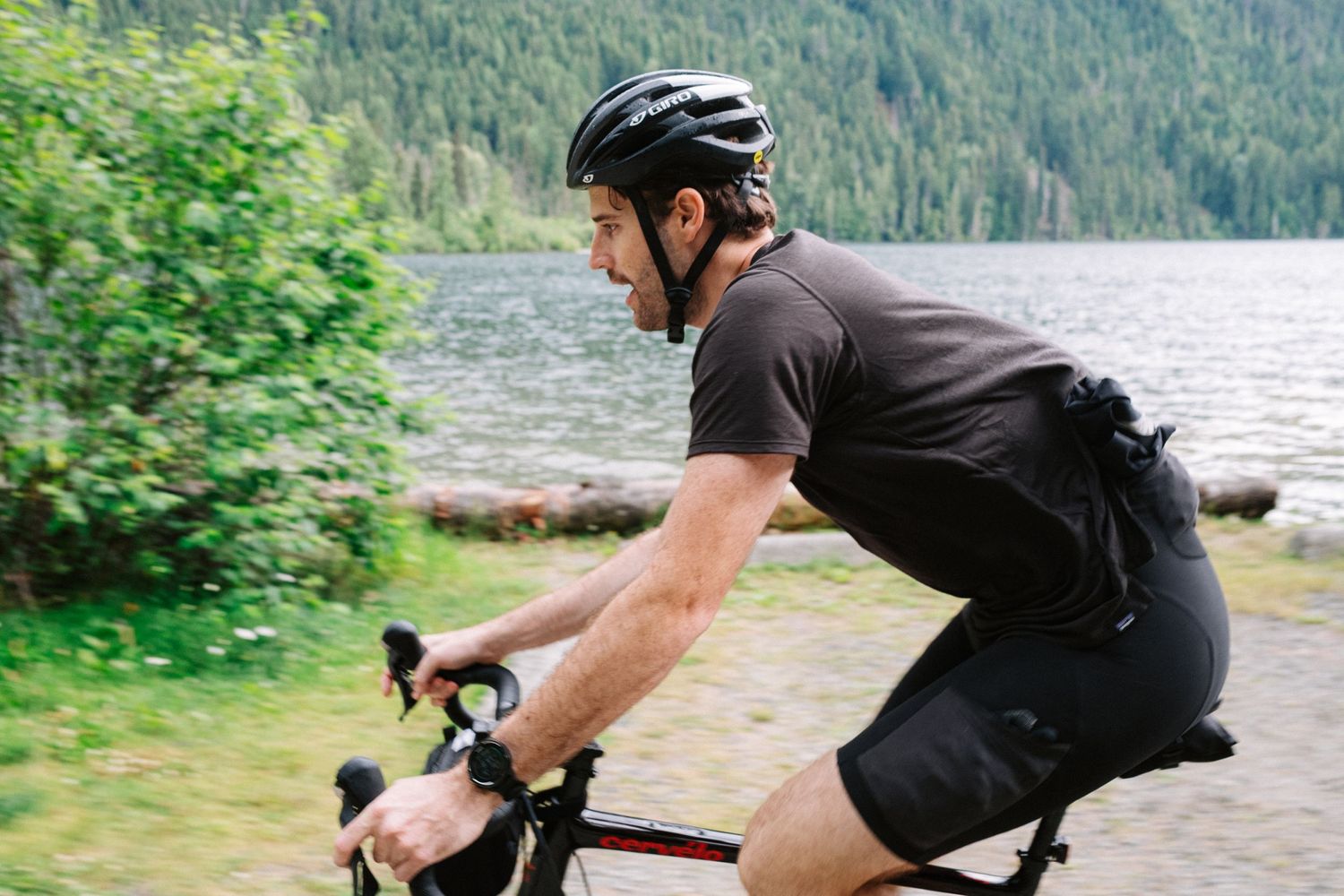 A cyclist in dark clothing rides along a scenic lakeside path with mountains and forest in the background.