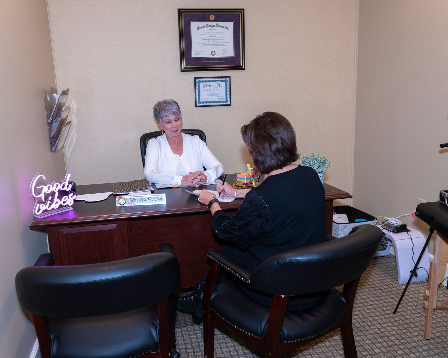 Professional woman consultant meeting with client at office desk with diploma on wall and Good Vibes neon sign.