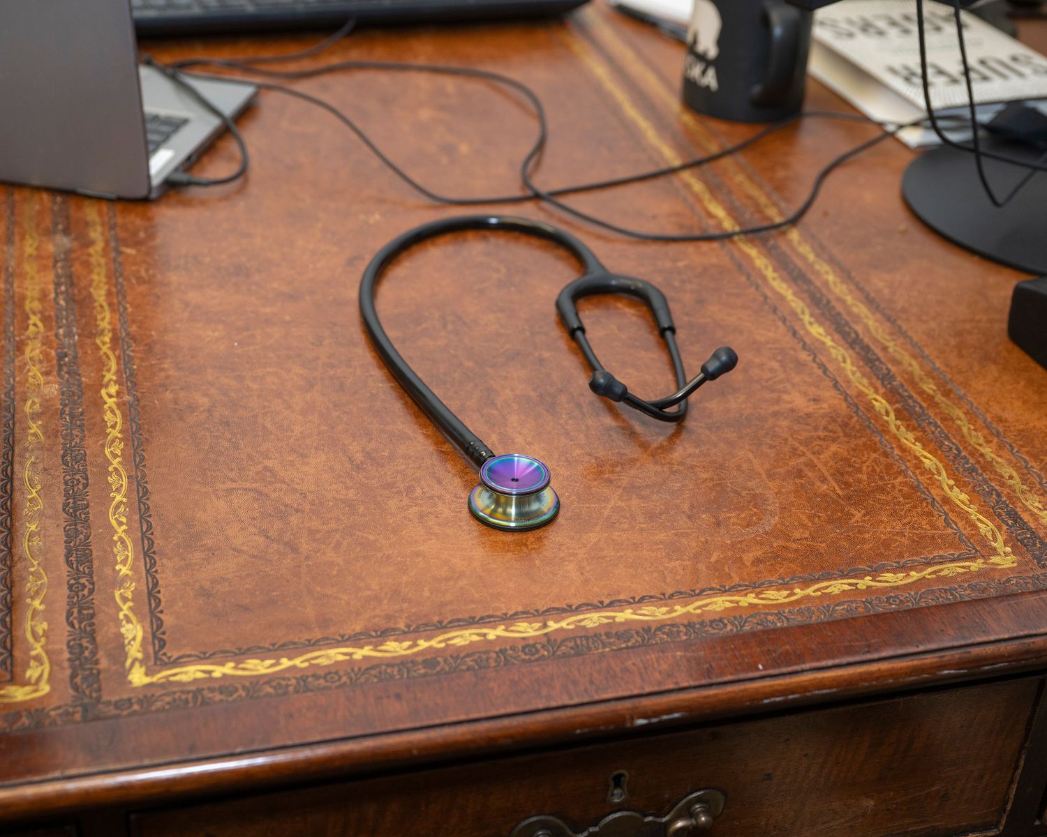 Black stethoscope with colorful chest piece resting on a vintage brown leather-top wooden desk.