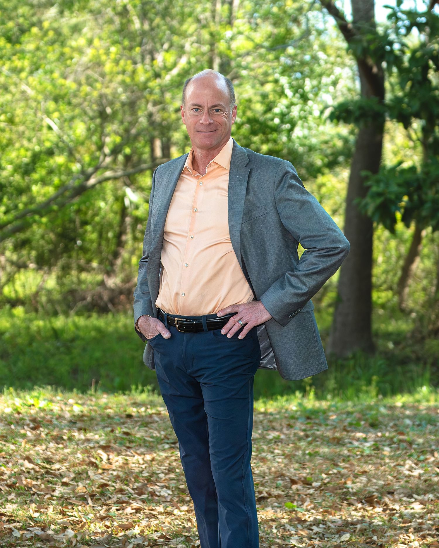 Professional man in gray blazer and peach shirt standing outdoors in a lush green park setting.