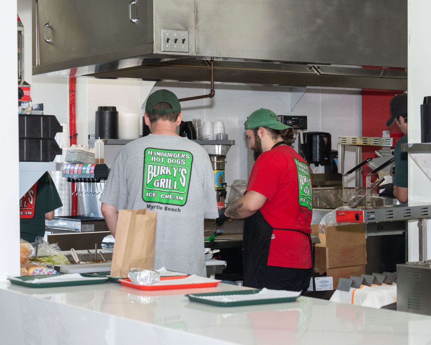 Two workers in green caps prepare food at a busy restaurant kitchen counter with cooking equipment behind them.
