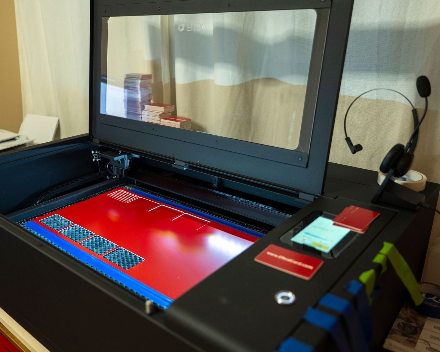 A laser engraver machine with red cutting bed open, displaying blue honeycomb tray, with headphones on desk nearby.