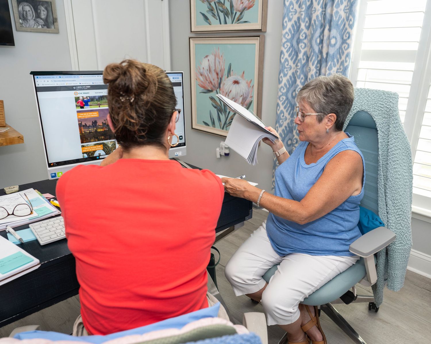 Two women reviewing documents together in a home office with a computer and coastal artwork on the wall.