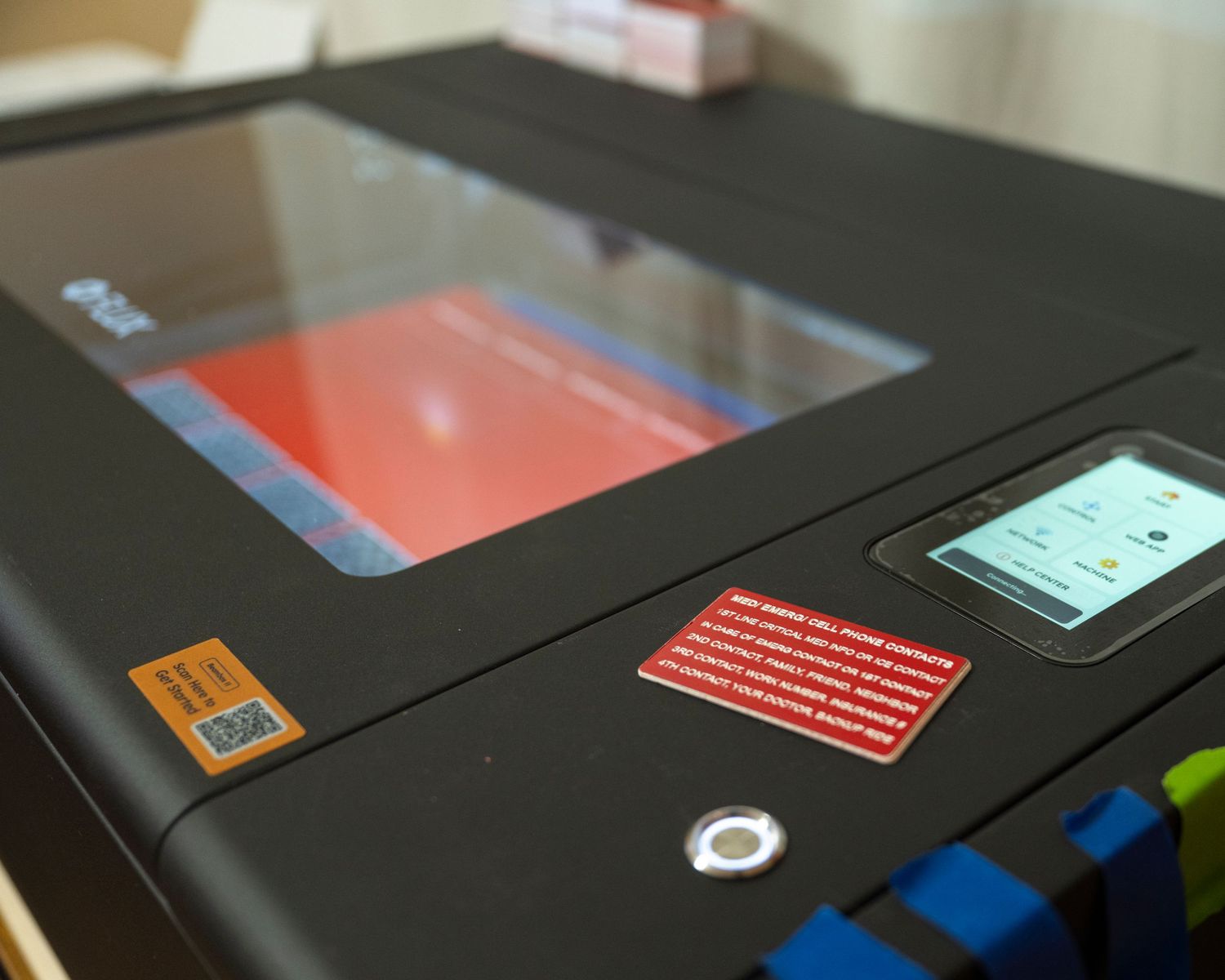Close-up of a black laser engraver machine with a red warning label and digital control panel display.