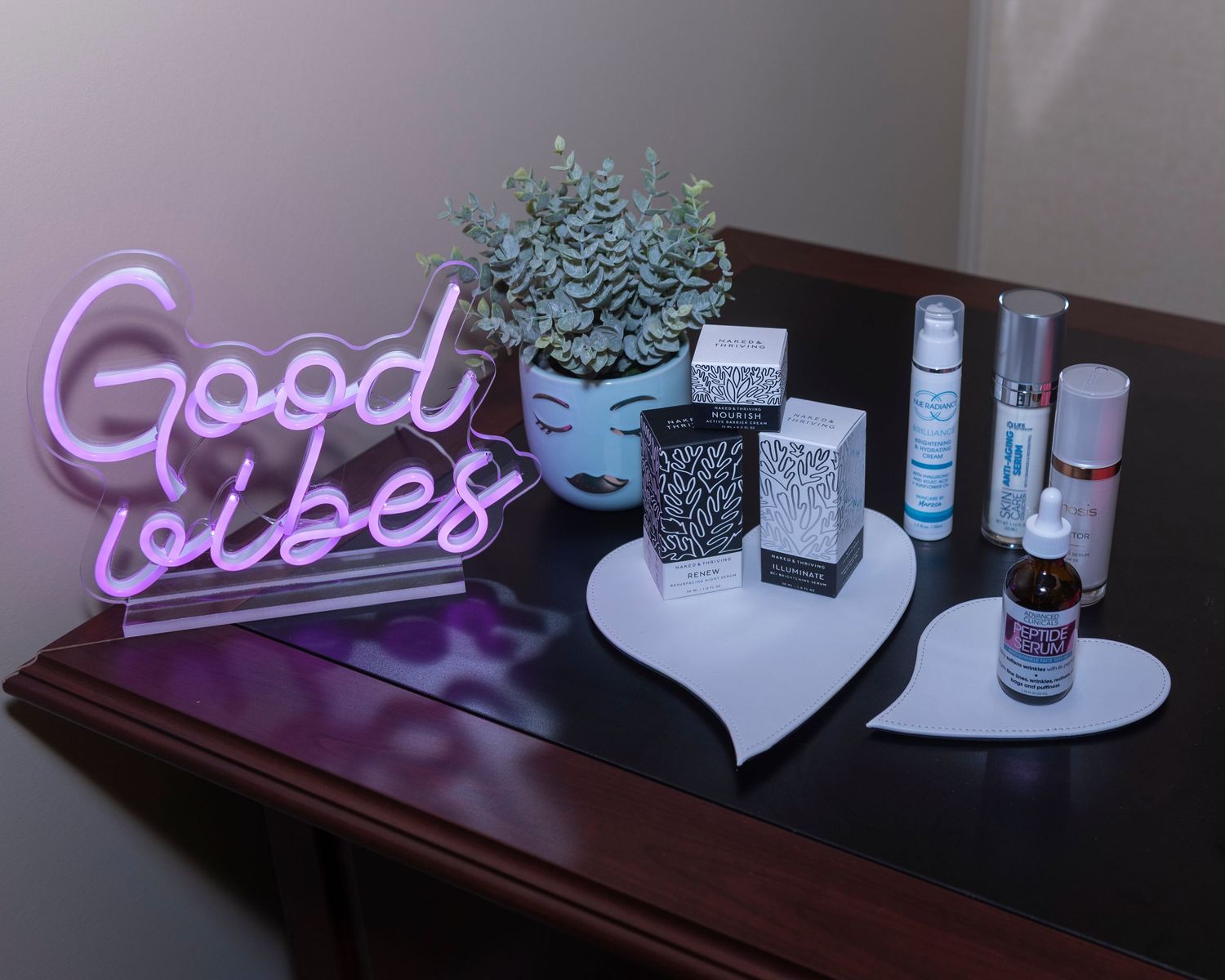 A wooden table displays a pink neon Good Vibes sign, a potted plant, skincare products, and a heart-shaped tray.