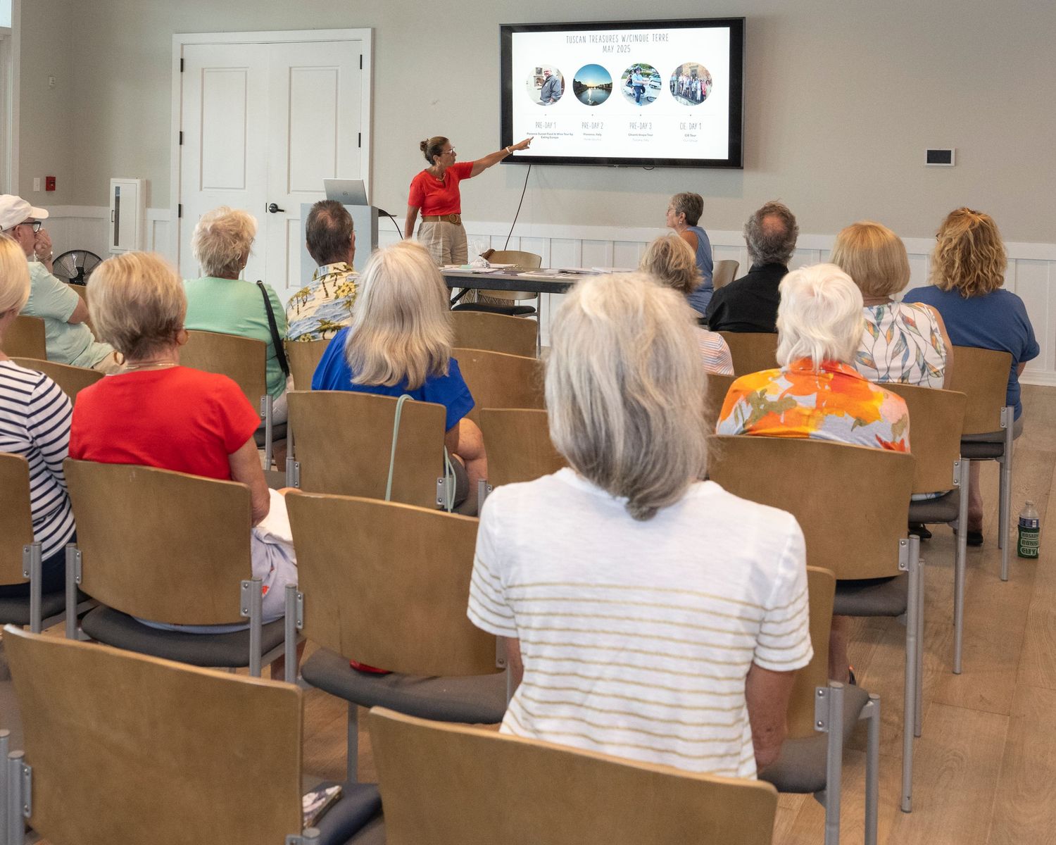 Elderly group attends a presentation with a speaker pointing at a large TV screen displaying colorful images.