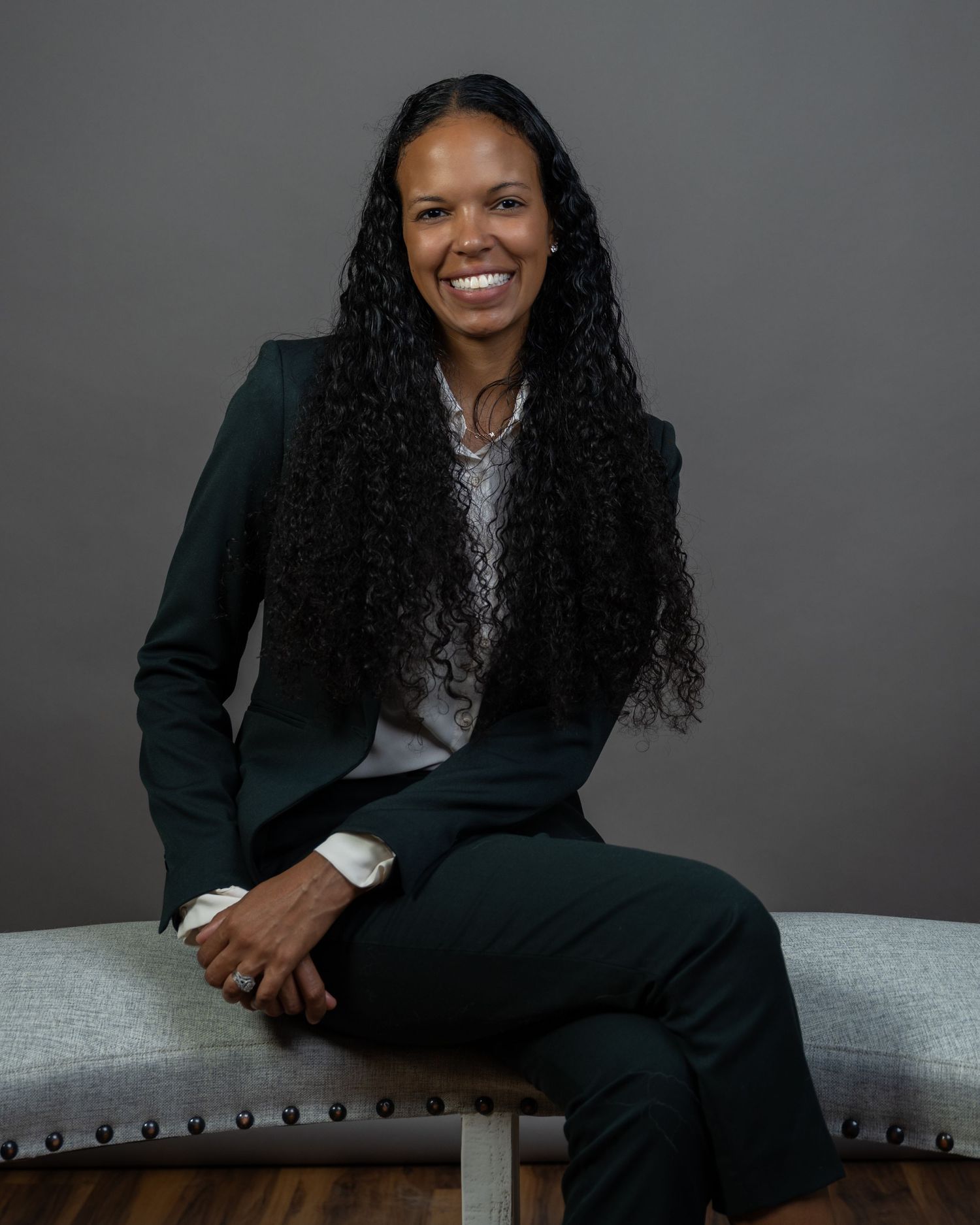 Professional woman in dark green suit with long curly hair smiling confidently while seated in a modern studio setting.
