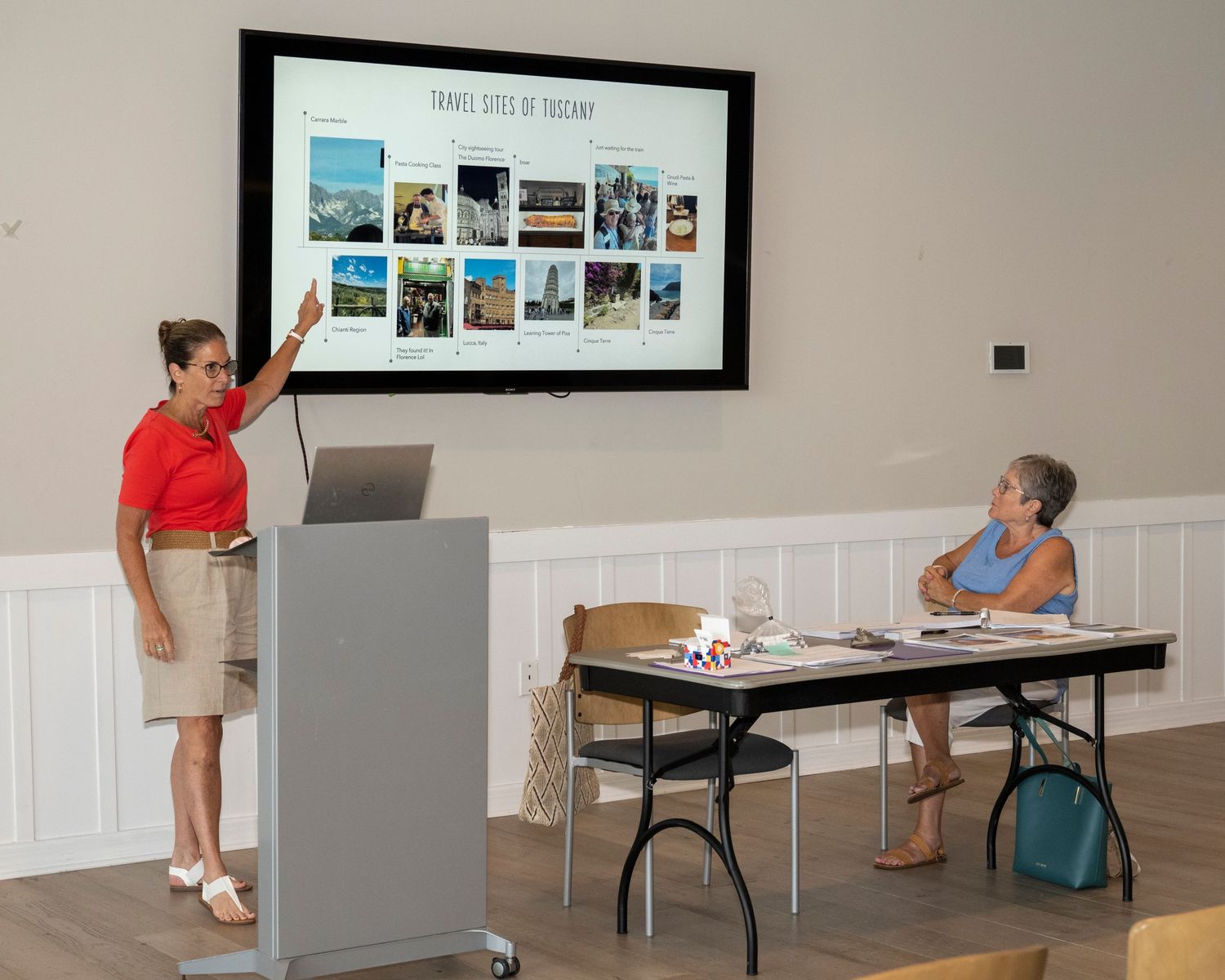 Woman presenting travel destinations on a large screen to an audience member seated at a table in a bright room.