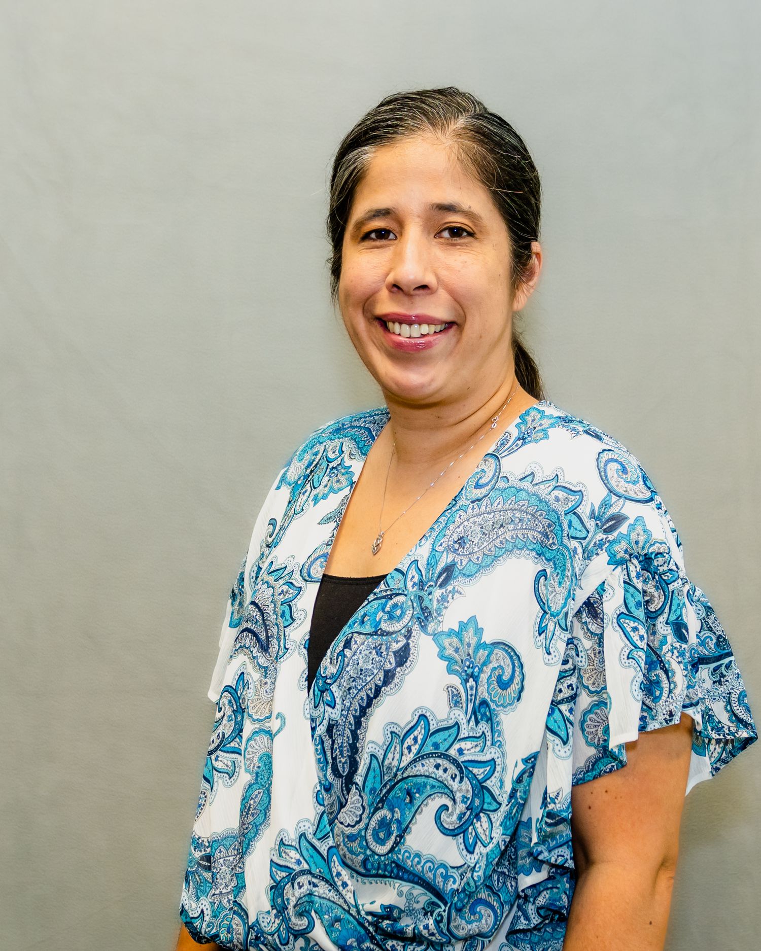 Smiling woman in blue and white paisley blouse poses for professional headshot photo against gray background.
