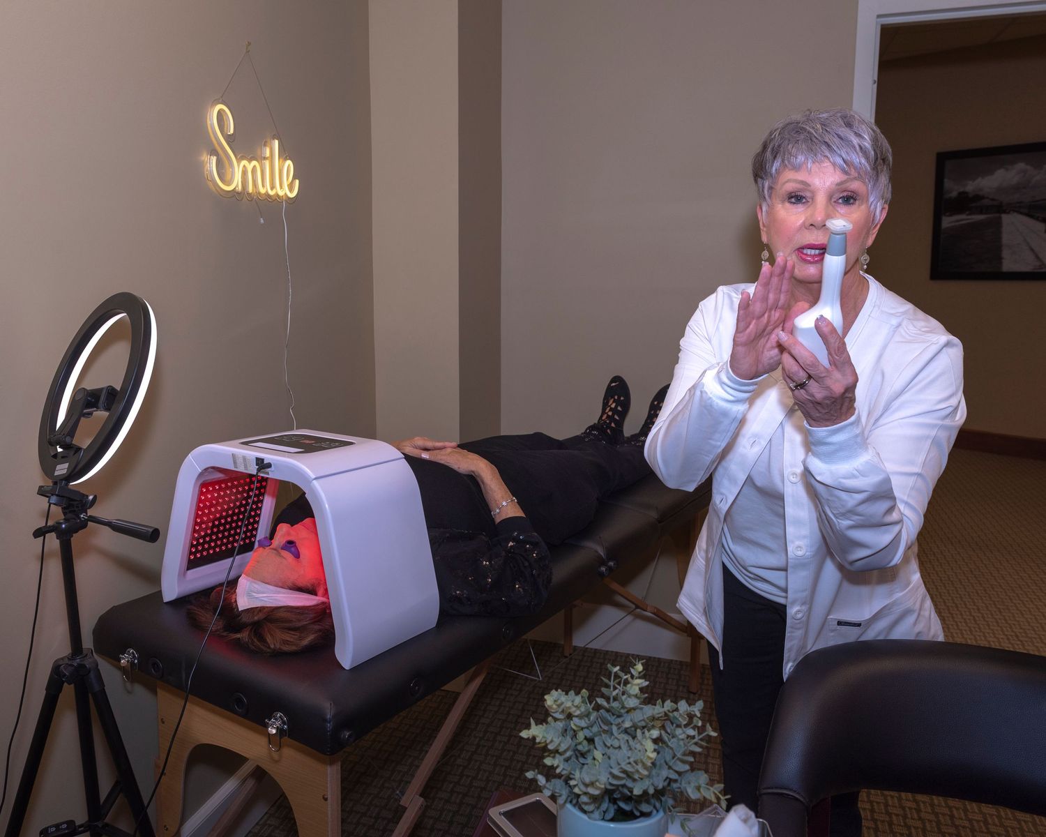 A woman in a white coat demonstrates a skincare device near a red light therapy bed with a ring light and Smile neon sign.