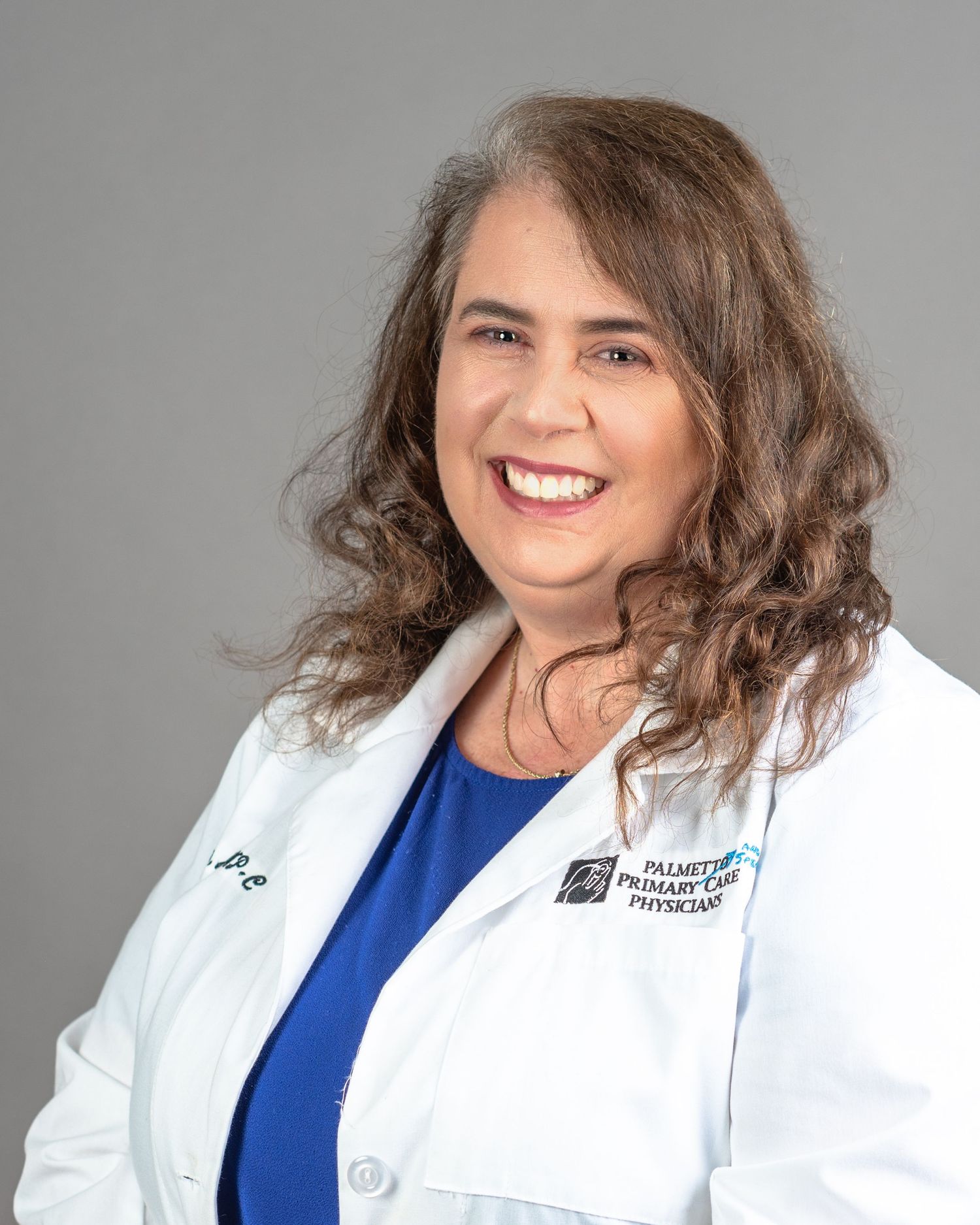 Female physician in white lab coat with Palmetto Primary Care logo smiling professionally against gray background.