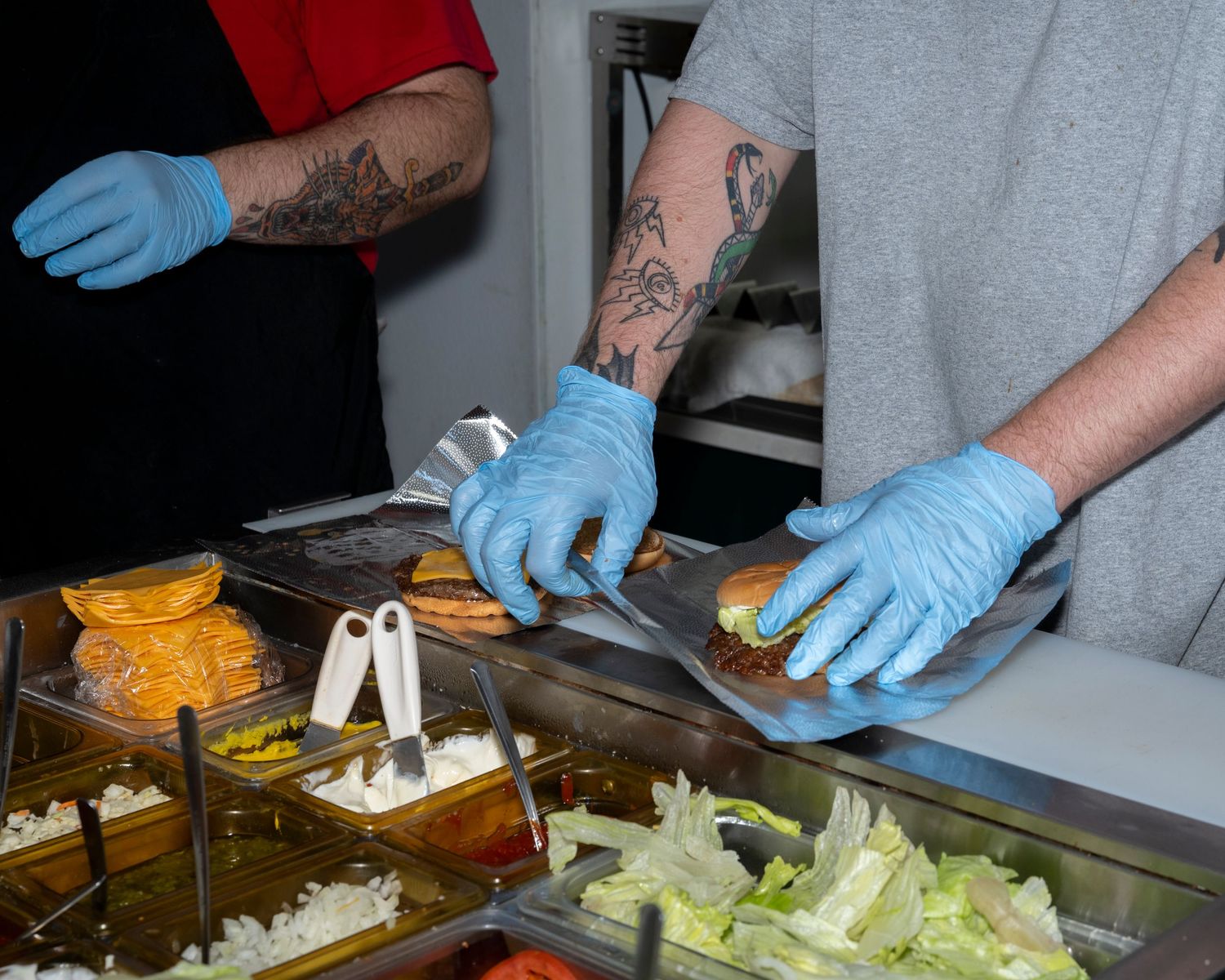 Tattooed food workers wearing blue gloves prepare fresh burgers at a sandwich counter with toppings.
