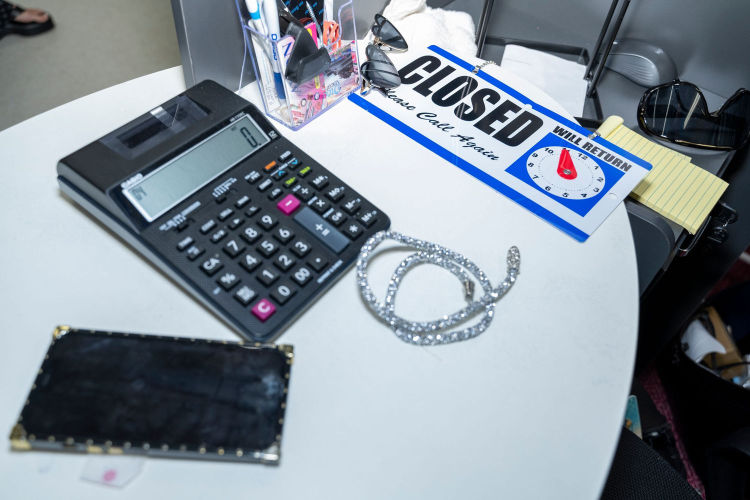 A desk with a calculator, chain, black wallet, and a closed sign with a timer on a white table.