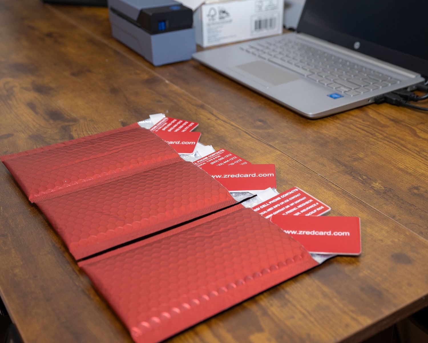 Red padded mailers with business cards on a wooden desk next to an HP laptop.