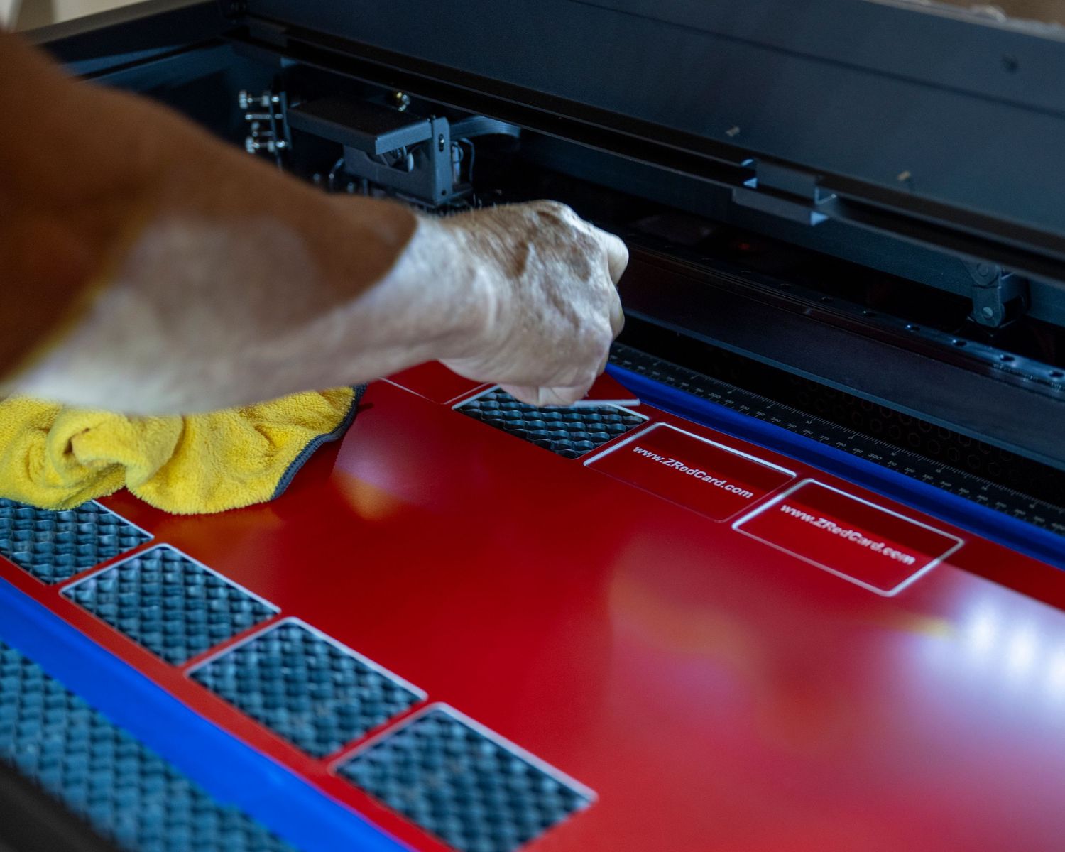 Hand polishing a red surface with a yellow cloth near a laser engraver with carbon fiber accents.