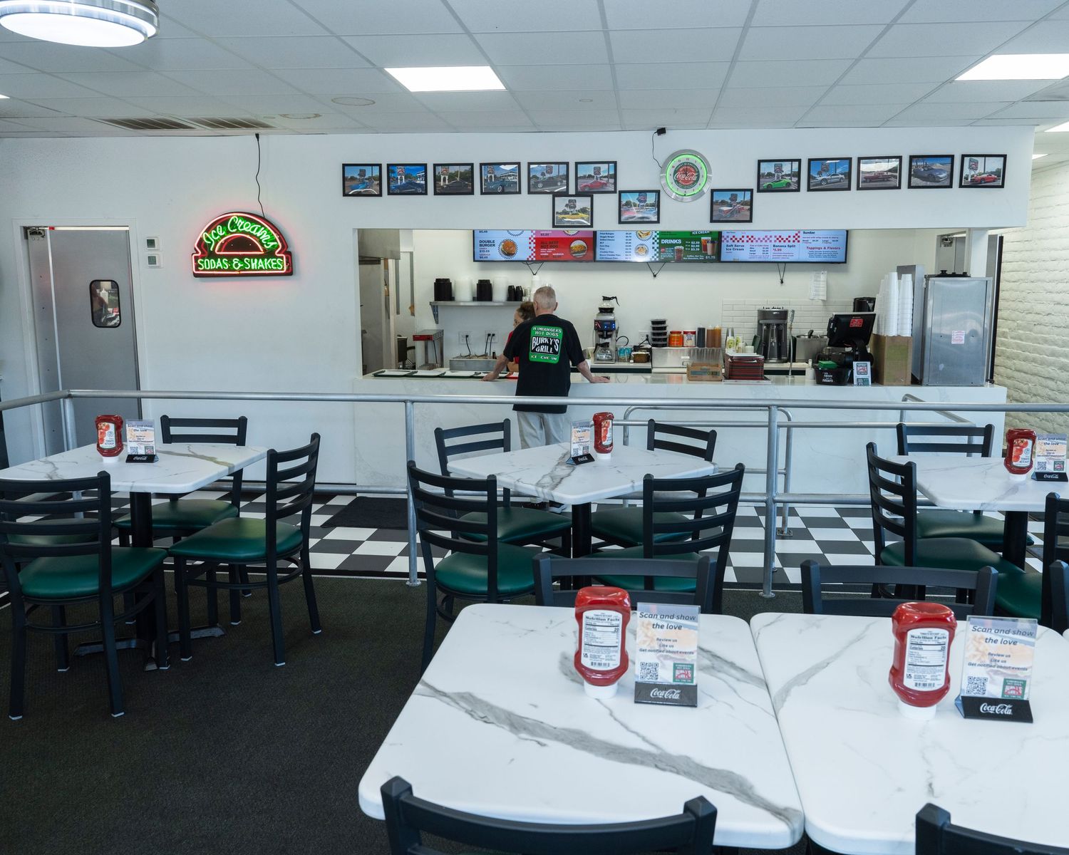 Interior of a casual diner with black chairs, white tables, checkered floors, and a service counter with a worker in green.