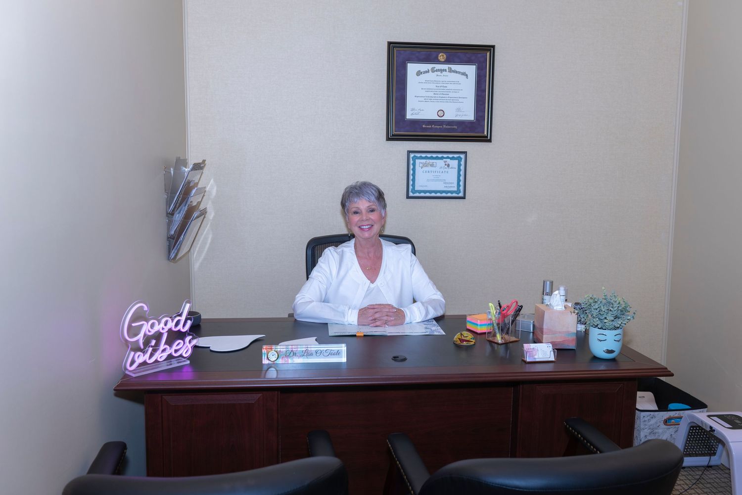 Professional woman in white coat smiling at office desk with diplomas on wall and Good Vibes neon sign.